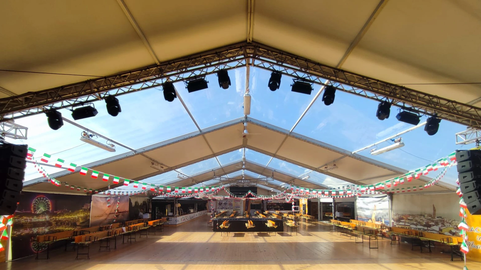 Interno di una grande tenda per eventi con soffitto trasparente, decorata con ghirlande rosse, bianche e verdi. Lungo i lati sono disposti lunghi tavoli e grandi altoparlanti neri sono posizionati nella parte anteriore. La luce del sole splendente filtra attraverso.