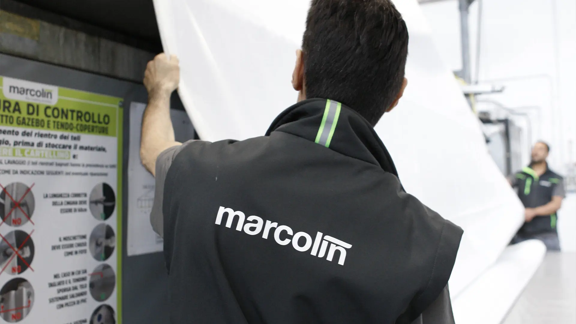Una persona che indossa un giubbotto "marcolin" sta ispezionando o sistemando un grande lenzuolo bianco in un ambiente industriale. Un'altra persona è visibile sullo sfondo. Ci sono cartelli di istruzioni di sicurezza su un muro vicino.