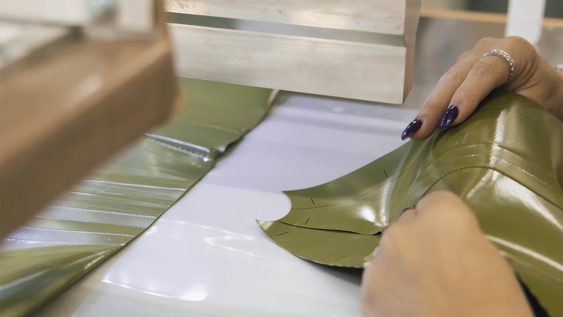 Mani con smalto scuro che tengono un tessuto verde, guidandolo attraverso una macchina da cucire. Contesto industriale con vista parziale della macchina sullo sfondo.
