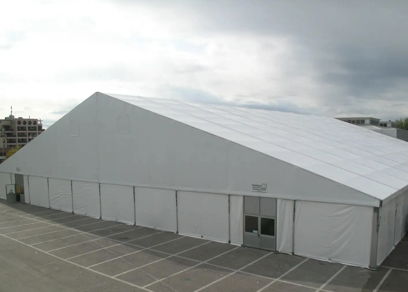 Una grande tenda bianca allestita in un parcheggio con una griglia di posti auto visibile. Il cielo è coperto e sullo sfondo si vedono degli edifici. La tenda ha i lati chiusi e alcune porte d'ingresso visibili.