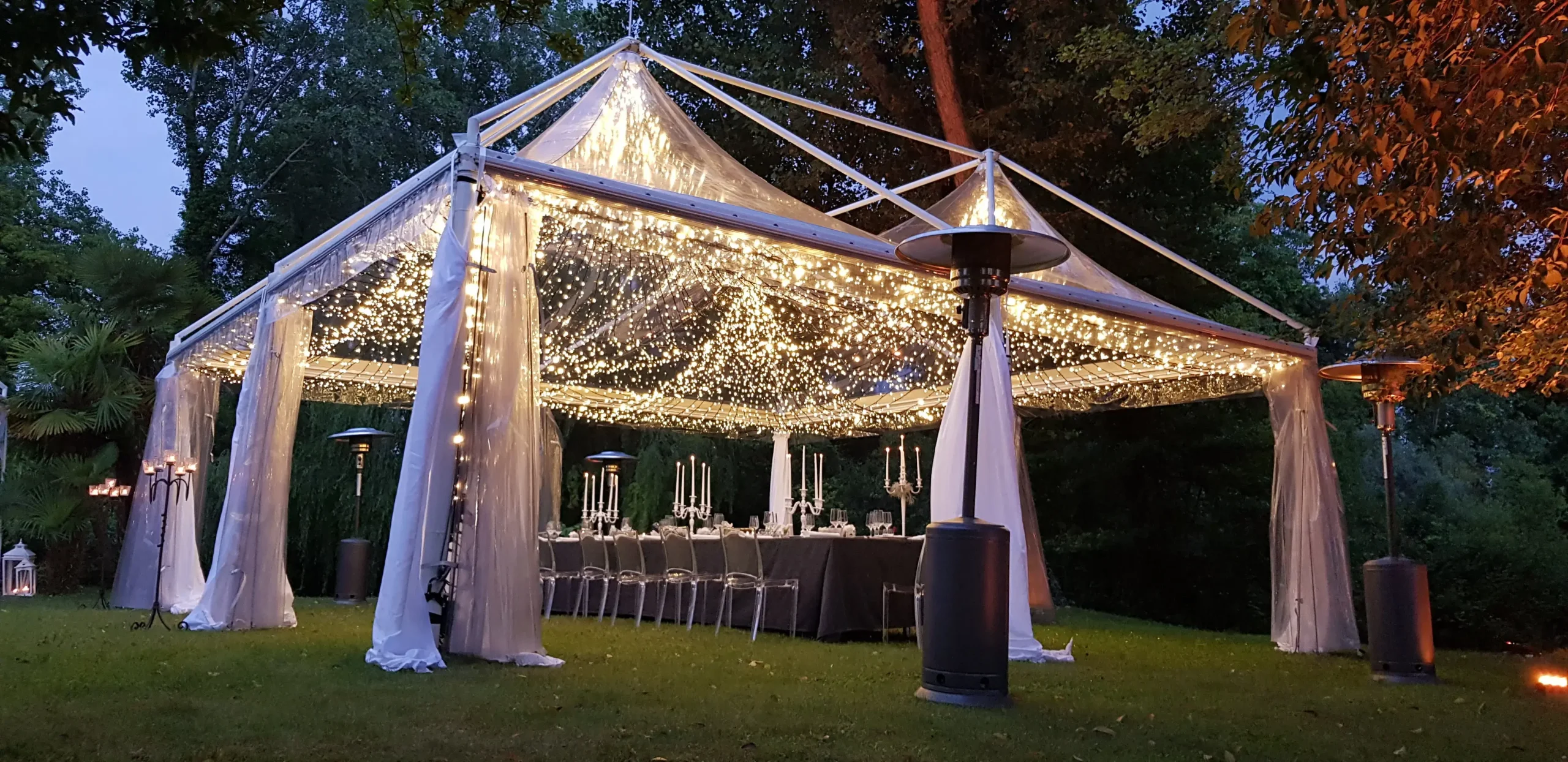 Una tenda trasparente illuminata da lucine viene allestita all'aperto al tramonto. All'interno, sotto il baldacchino, è allestito un lungo tavolo con eleganti tovaglie e alti candelabri. L'area circostante è lussureggiante e verdeggiante.