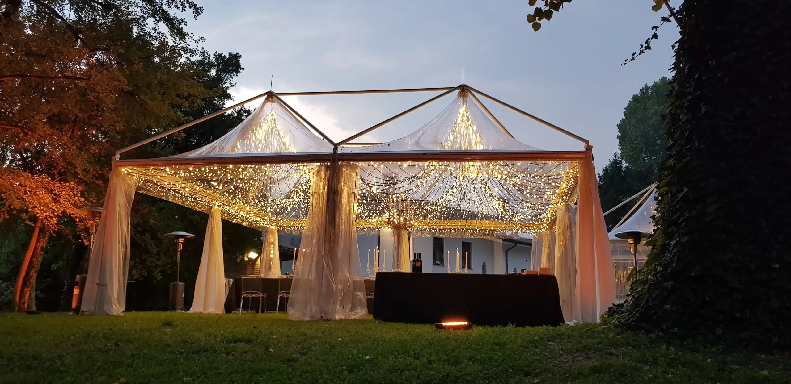 Una tenda da matrimonio all'aperto splendidamente illuminata con lucine a drappeggio, situata su un prato erboso. La tenda è incorniciata da alberi e presenta tende bianche trasparenti, che creano un'atmosfera romantica.