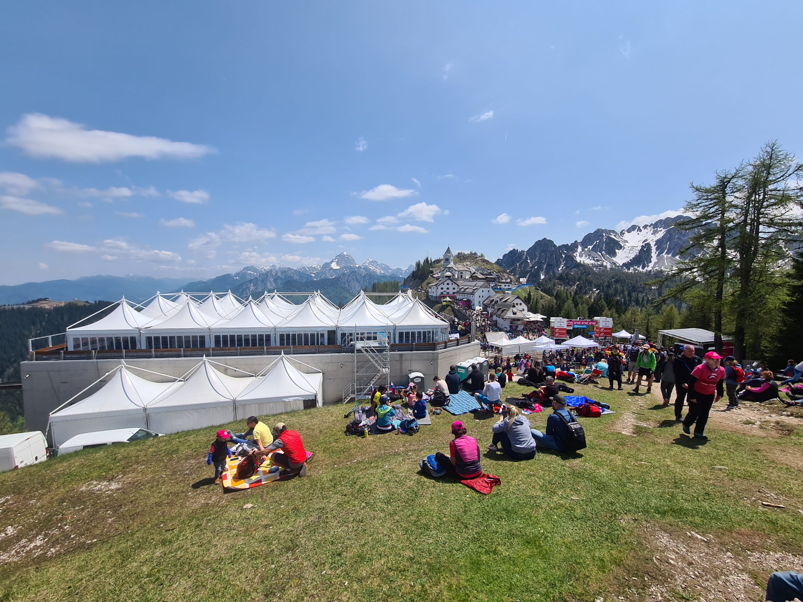 La gente si siede e si rilassa su un pendio erboso vicino a tende bianche con montagne e neve sullo sfondo, sotto un cielo azzurro con nuvole sparse. Alcuni si radunano vicino a colorati stand per eventi.