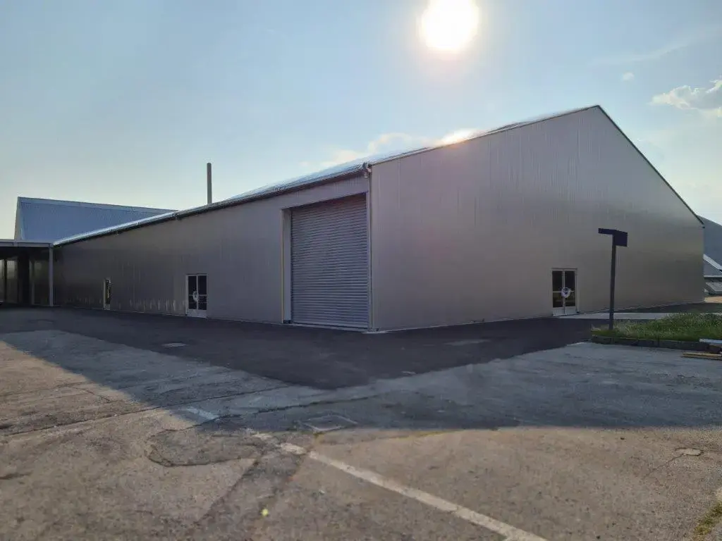 Grande magazzino industriale con esterno in metallo argentato e portone avvolgibile chiuso. Il sole splende in alto, proiettando ombre. L'edificio è circondato da terreno pavimentato e prato. Sullo sfondo, un cielo azzurro e terso.