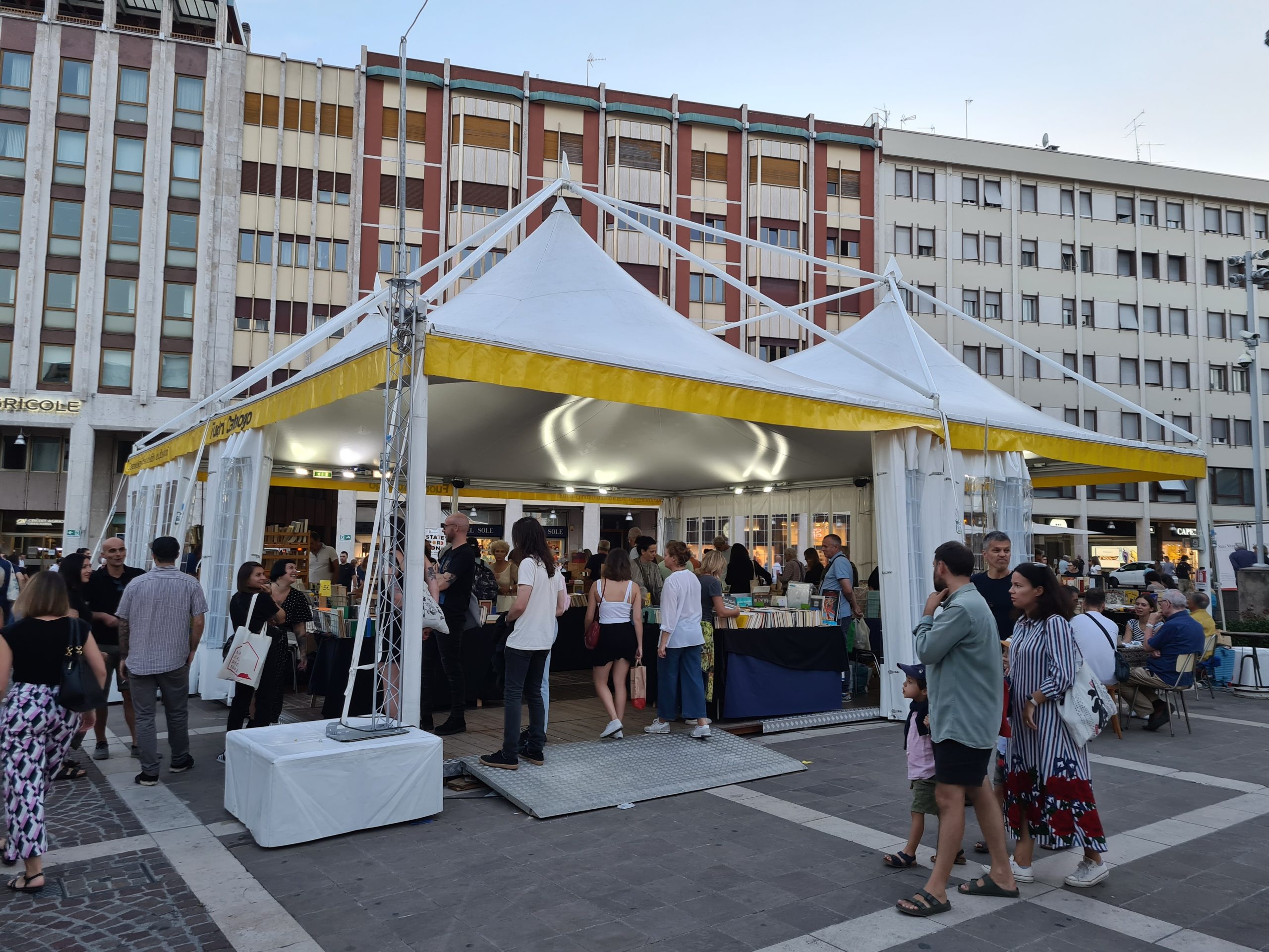 Una tenda bianca con bordi gialli è allestita in una piazza cittadina, circondata da persone che sfogliano libri e chiacchierano. Sullo sfondo si vedono alti edifici e la scena sembra essere quella di un evento all'aperto o di una fiera.