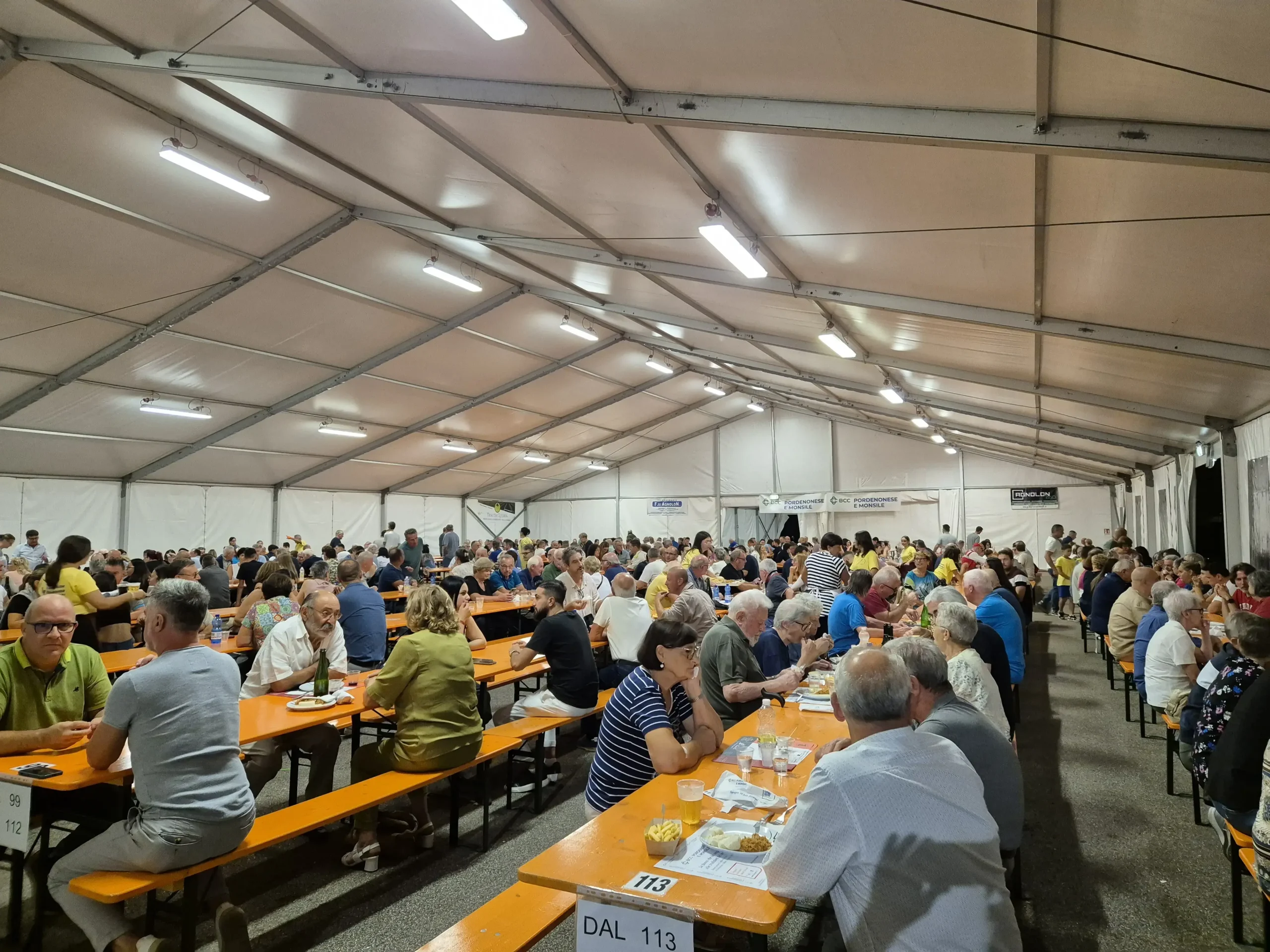 Un folto gruppo di persone sedute a lunghi tavoli condivisi sotto un'ampia tenda bianca. Sembra che stiano mangiando e socializzando in un'atmosfera vivace, circondati da un arredamento semplice e da luci soffuse.