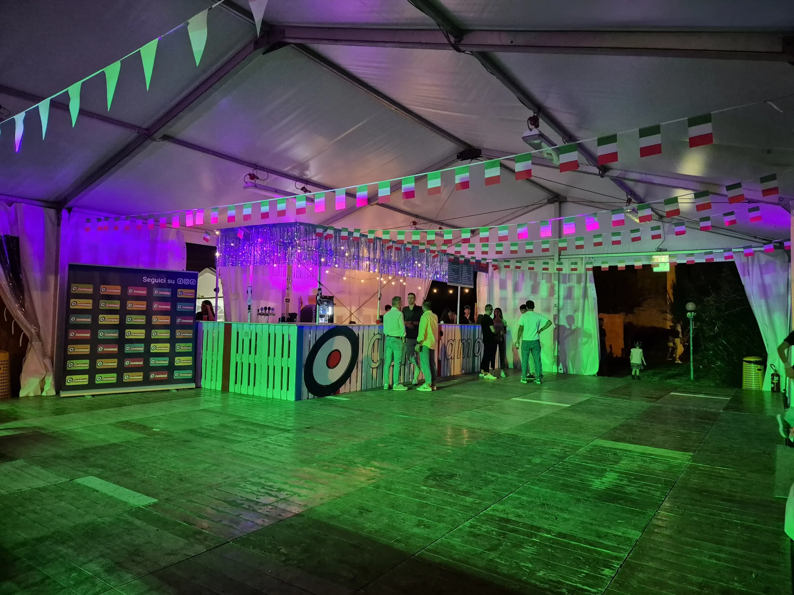 Un tendone per eventi illuminato a giorno con luci e decorazioni colorate. La gente si raduna attorno al bar, decorato con bandierine e un bersaglio circolare. Il pavimento è in legno e l'atmosfera è festosa.
