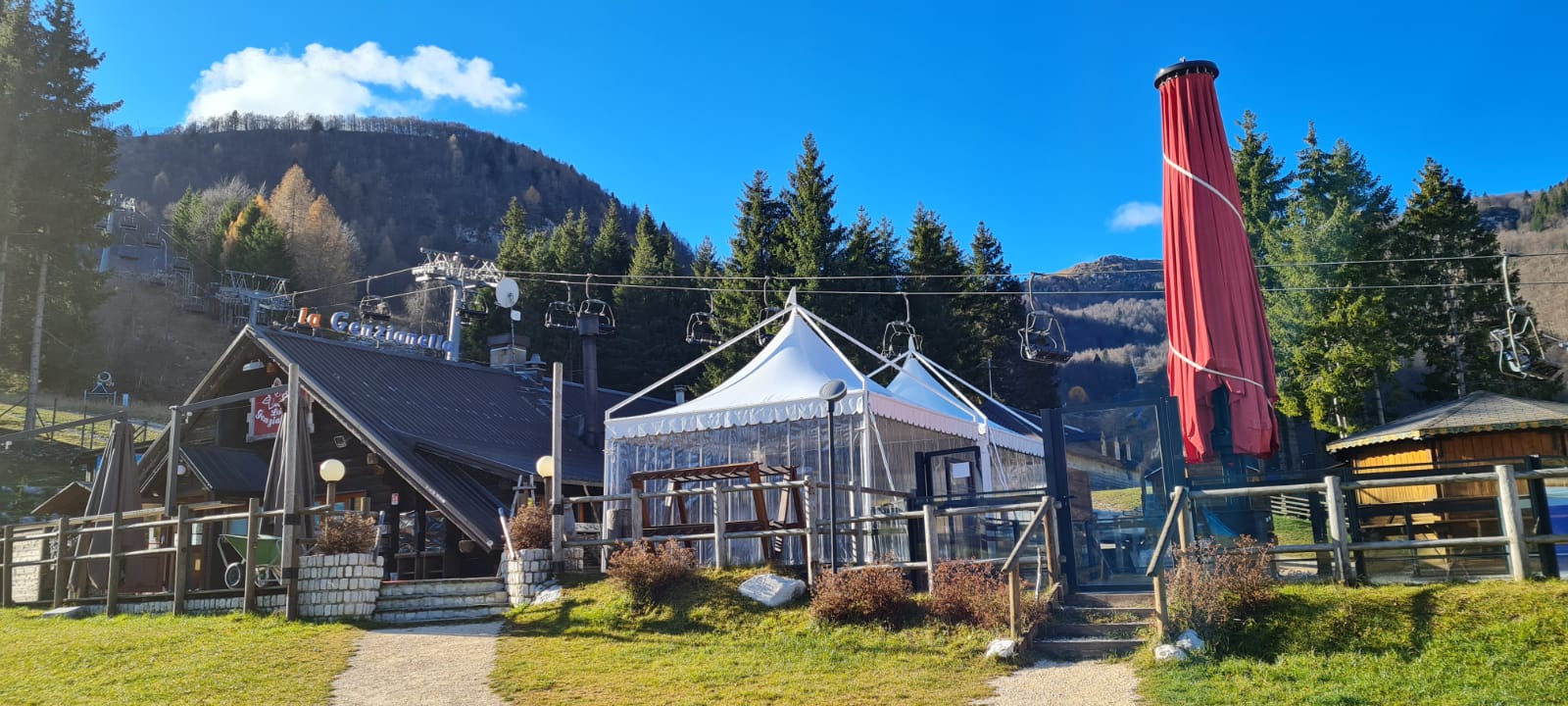 Un rustico ristorante di montagna con tetto spiovente e terrazza vetrata si erge tra pini e colline. Fuori c'è un grande ombrellone rosso chiuso, e gli impianti di risalita vuoti incombono sopra di noi. Il cielo è limpido e azzurro.