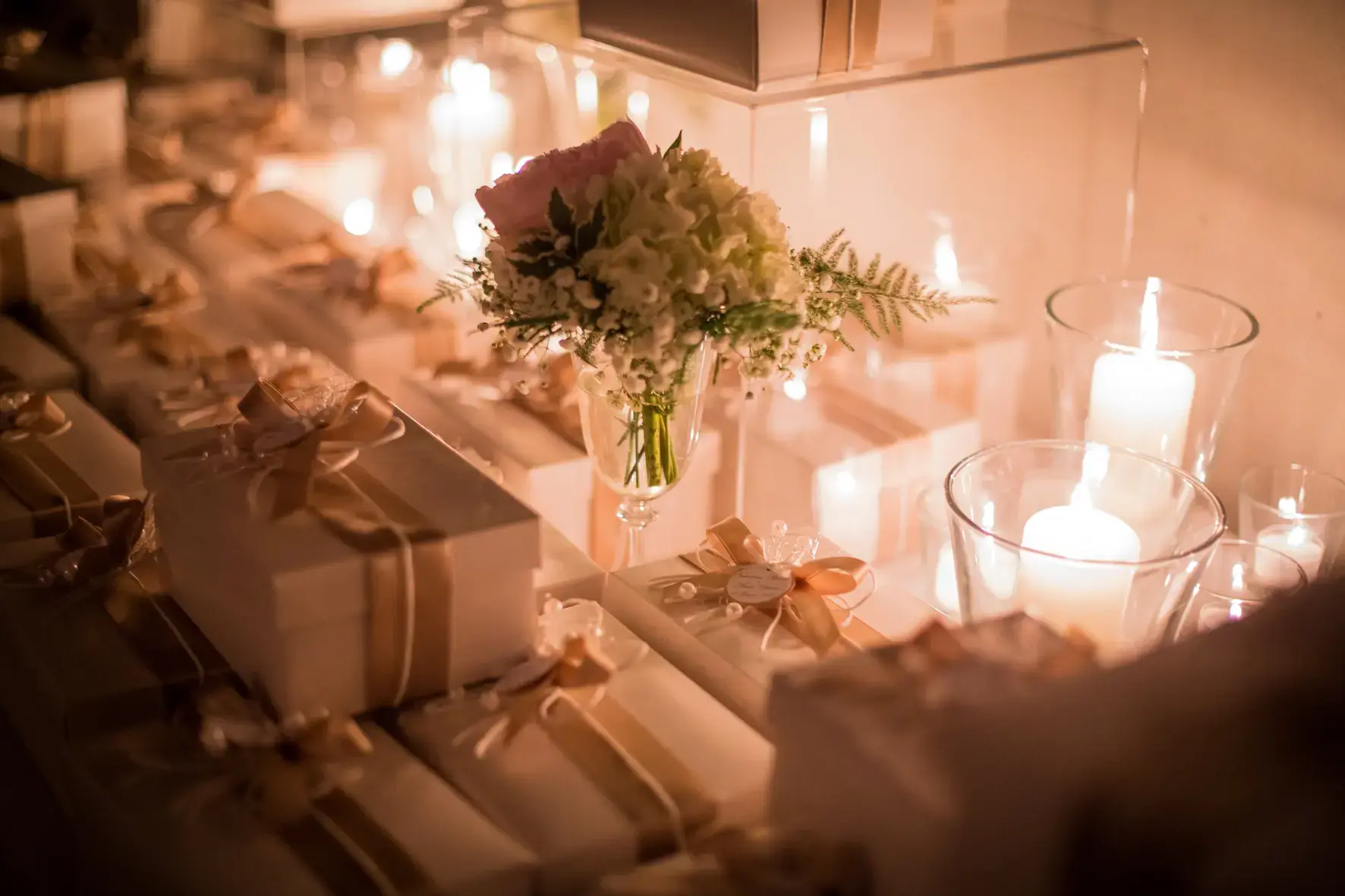 Un tavolo decorato con regali elegantemente confezionati in bianco e oro, con al centro un bouquet di fiori bianchi e rosa in un vaso di vetro. La luce soffusa delle candele illumina la scena, creando un'atmosfera calda e romantica.