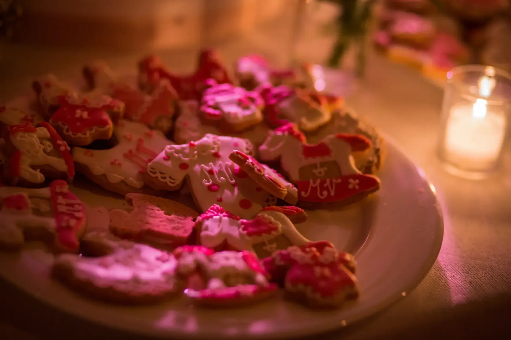 Un piatto pieno di vari biscotti rosa e bianchi decorati con glassa colorata. I biscotti hanno la forma di abiti e altri indumenti. Una candela accesa in un portacandele di vetro proietta una luce calda sulla scena.