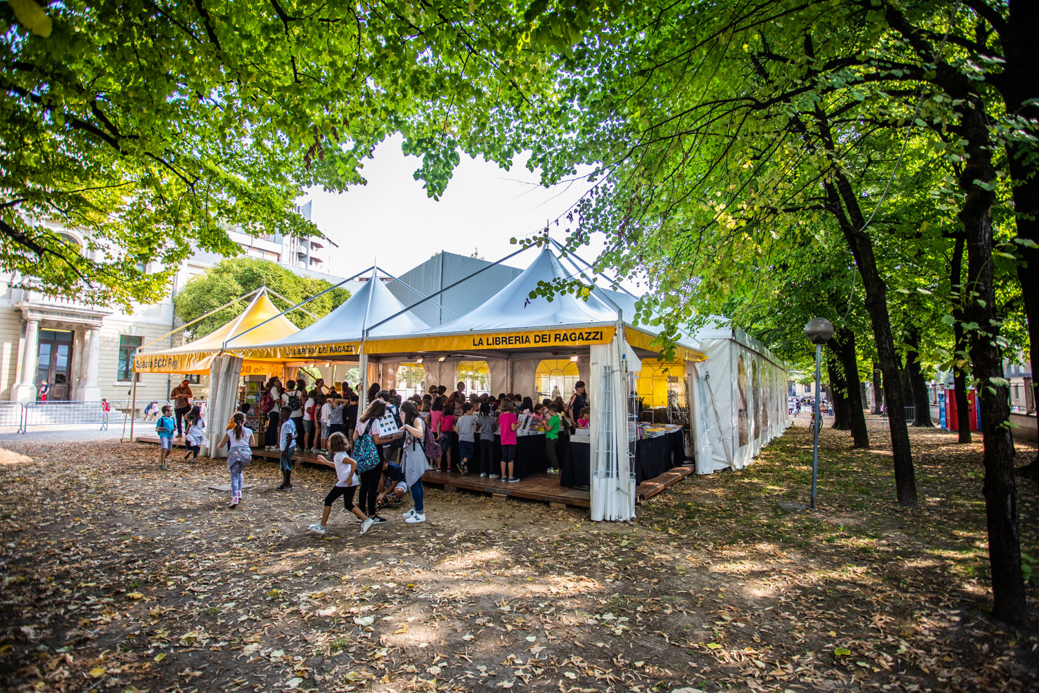 Un gruppo di persone, tra cui bambini, si riunisce sotto una tenda bianca e gialla con la scritta "La Libreria dei Ragazzi" in un parco circondato da alberi e foglie cadute.
