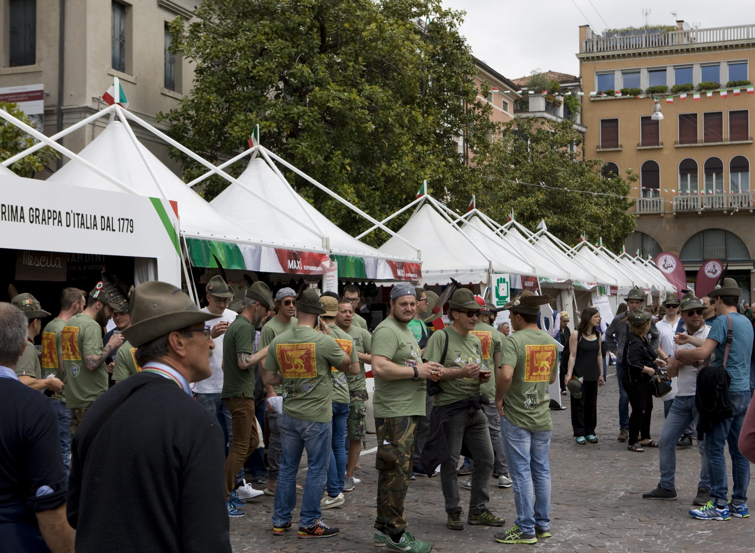Un gruppo di persone con camicie e cappelli verdi si raduna davanti alle tende bianche dei venditori ambulanti durante un evento all'aperto nella piazza di una città europea, con vecchi edifici e alberi sullo sfondo.
