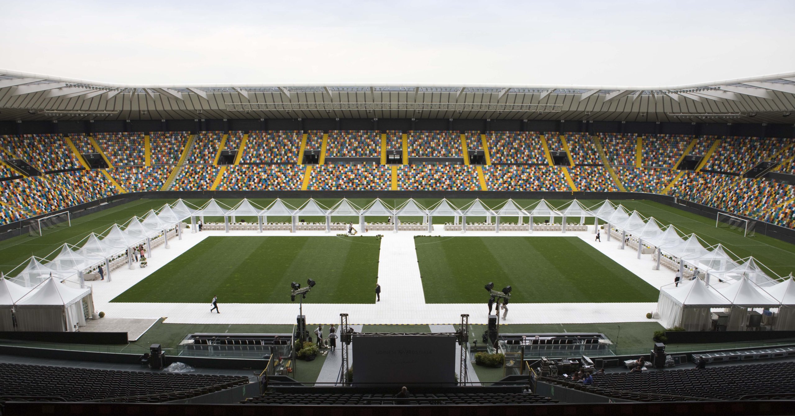 Un grande stadio di calcio con sedili multicolori e un campo verde, con file di tende bianche allestite al centro, probabilmente per un evento. Diverse persone camminano e lavorano vicino alle tende.