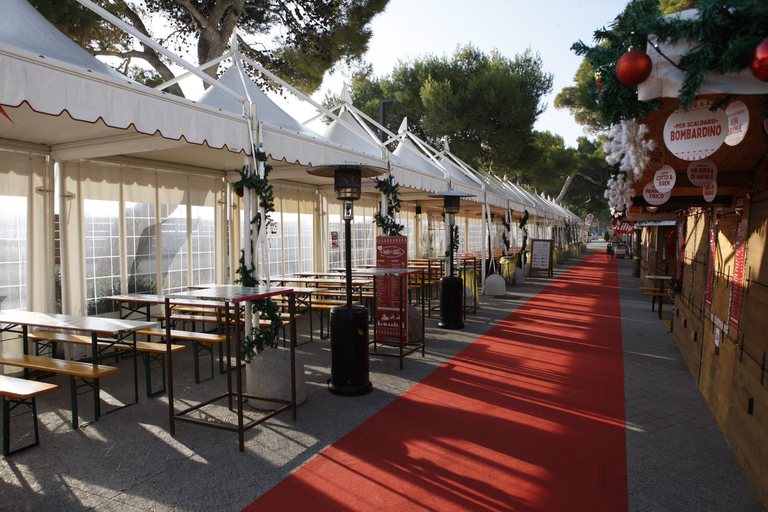 Una lunga tenda esterna con decorazioni festive e un tappeto rosso, file di tavoli e panche in legno da un lato e bancarelle in legno con decorazioni natalizie dall'altro, sotto gli alberi e con la luce del giorno che filtra attraverso.