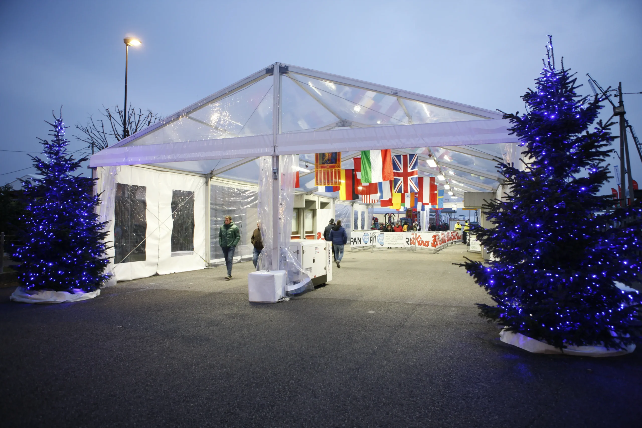 Un'area esterna festosa con una tenda trasparente decorata con bandiere di vari paesi. Due alberi di Natale illuminati con luci blu si trovano ai lati della tenda. Diverse persone passeggiano nelle vicinanze sotto un cielo nuvoloso.