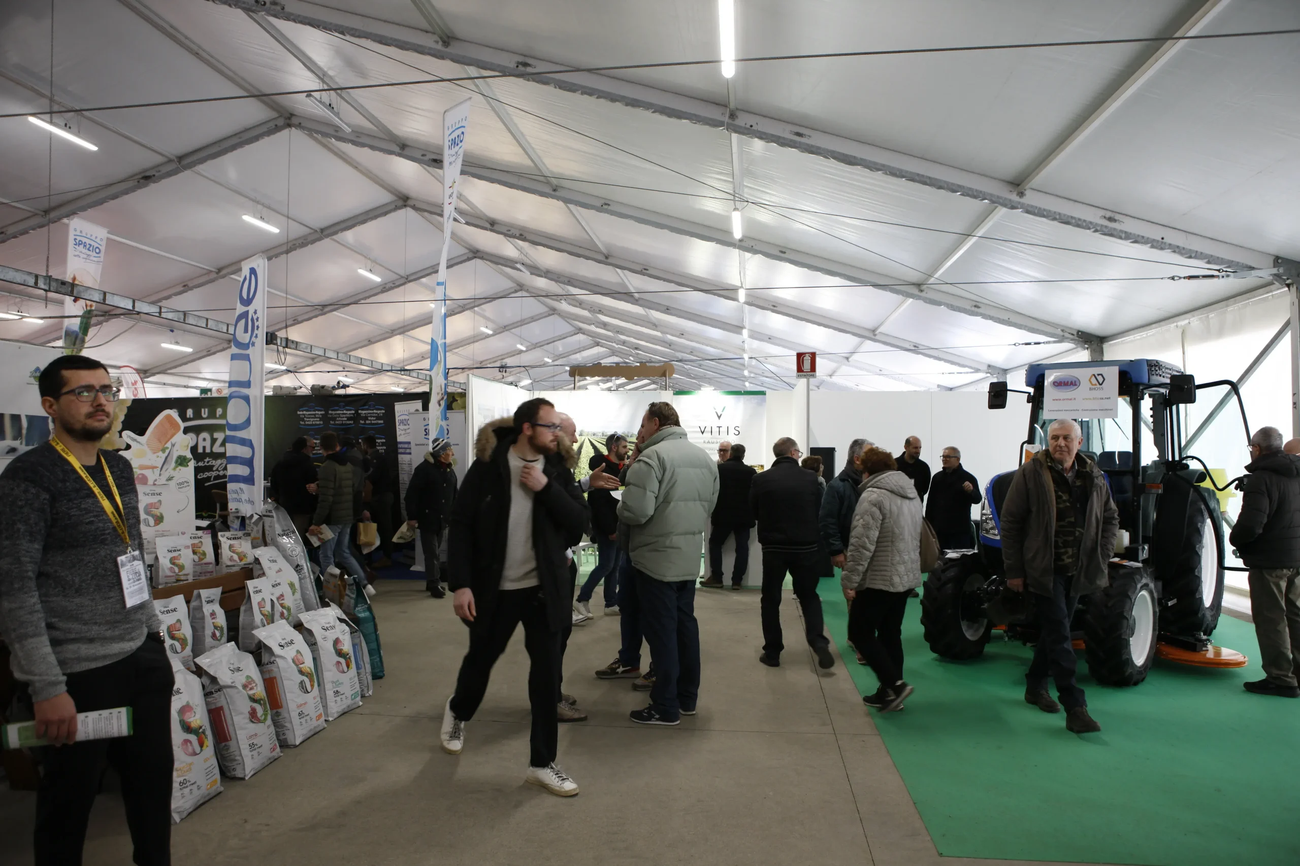 Persone che passeggiano e curiosano in una fiera commerciale sotto un grande tendone. Ci sono diverse esposizioni, tra cui prodotti agricoli e un trattore blu. Alcuni partecipanti interagiscono con gli espositori. L'atmosfera è vivace e informale.