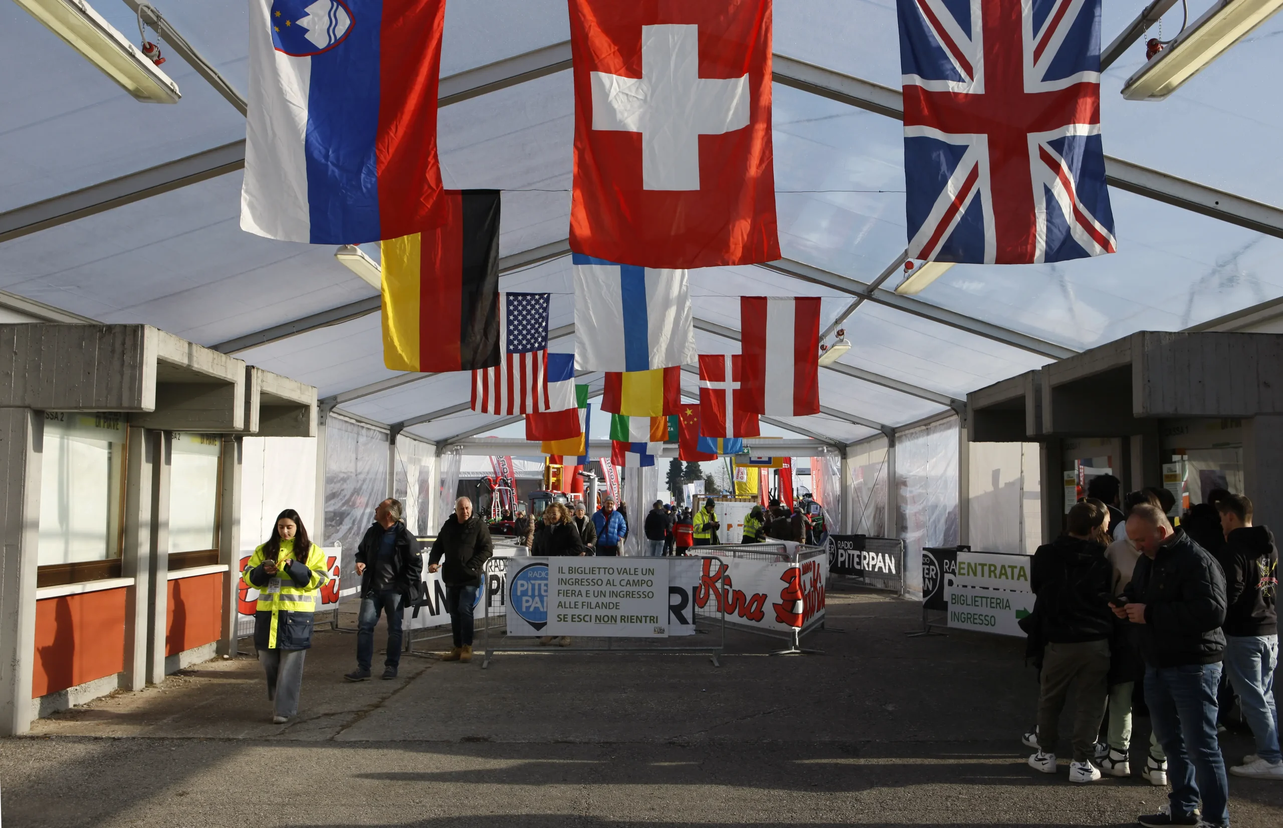 Un gruppo di persone attraversa una tenda decorata con bandiere di vari paesi, tra cui Slovenia, Svizzera e Regno Unito. All'ingresso sono visibili cartelli e recinzioni, che indicano un evento o un raduno. L'atmosfera è vivace e organizzata.