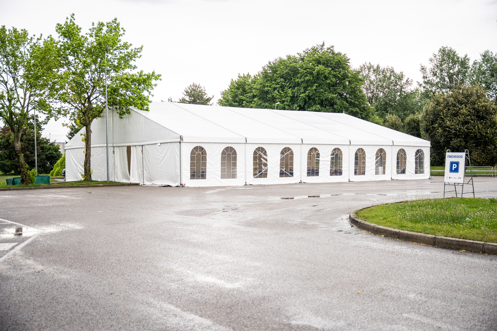 Un grande tendone bianco per eventi con finestre ad arco è allestito su un'area pavimentata e bagnata, vicino a un prato erboso e ad alcuni alberi. Un cartello con una "P" indica il parcheggio nelle vicinanze. Il cielo è coperto.