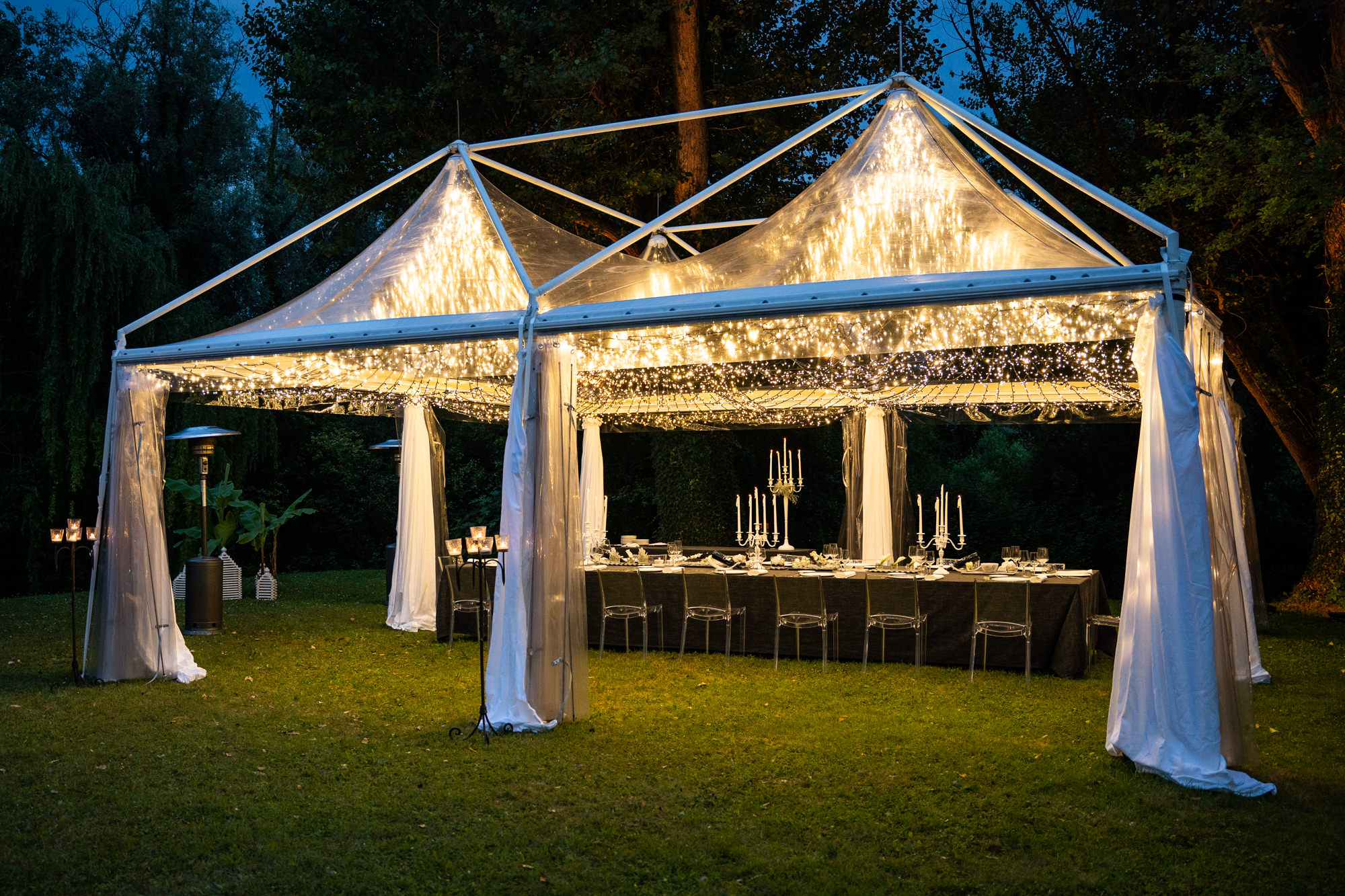 Un grande tavolo all'aperto apparecchiato per la cena sotto una tettoia trasparente decorata con lucine sospese e tende bianche trasparenti, creando un'atmosfera calda ed elegante sullo sfondo degli alberi al tramonto.