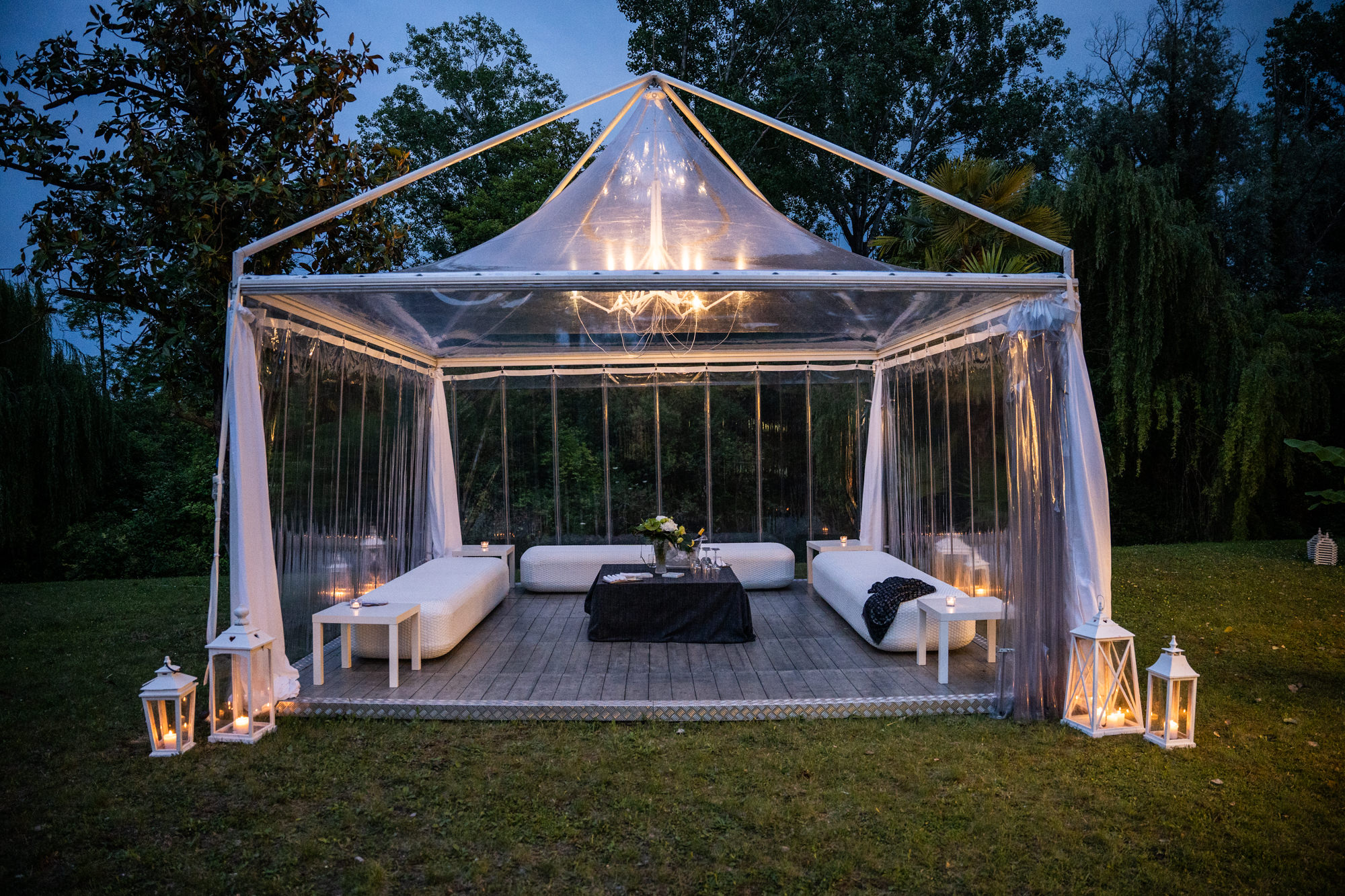Una tenda esterna trasparente con divani bianchi, un tavolo ricoperto di nero, un centrotavola floreale e lanterne accese, posta su una piattaforma di legno in un'area erbosa circondata da alberi al tramonto.