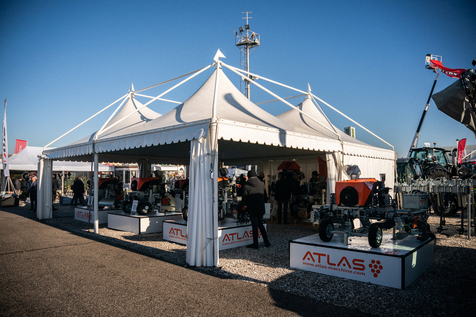 Una tenda bianca espone diversi macchinari su piattaforme con la scritta "ATLAS" durante un evento all'aperto. Le persone osservano le attrezzature sotto un cielo azzurro e limpido.