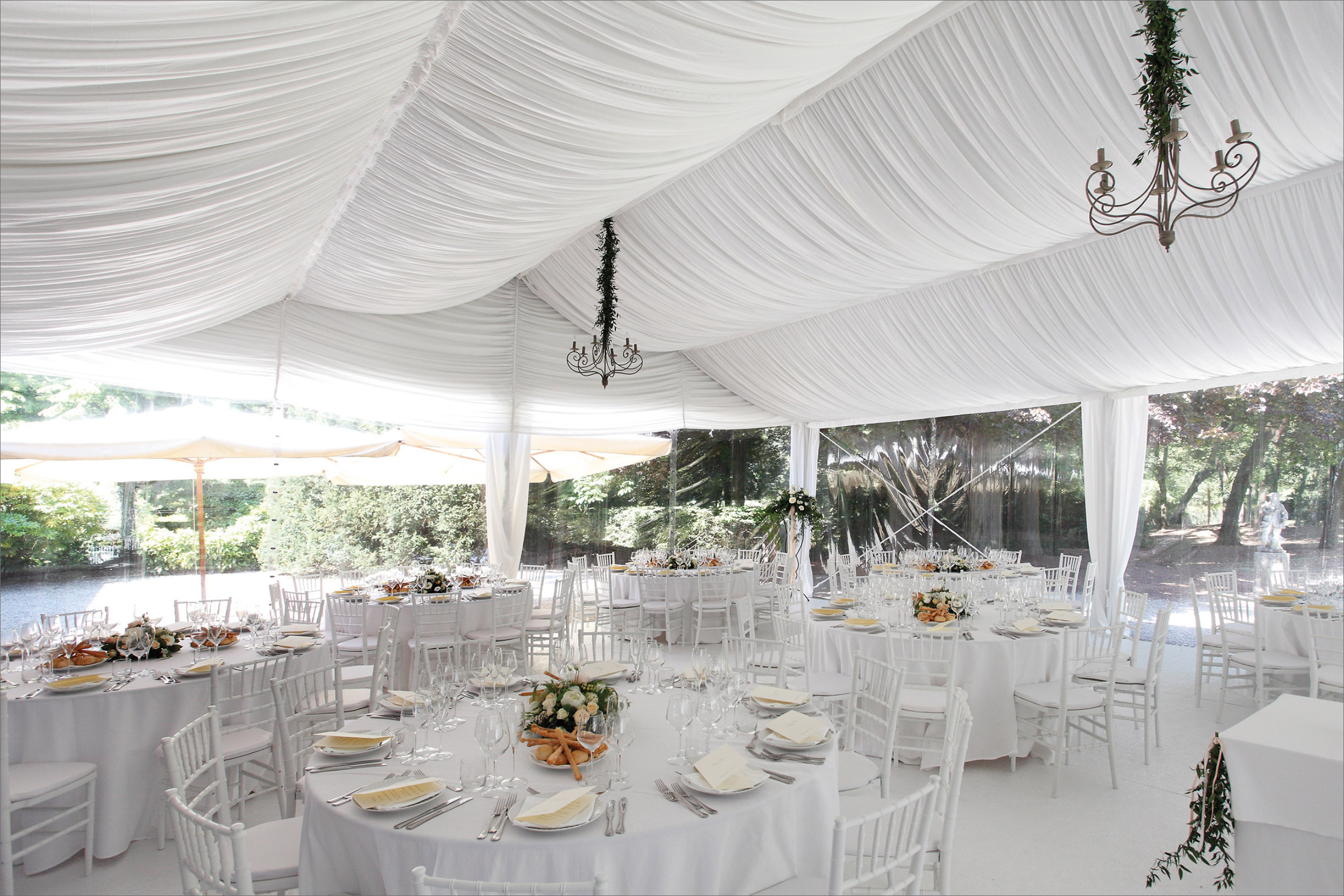 Un'elegante tenda bianca allestita per un evento, con tavoli rotondi coperti da tovaglie bianche, sedie bianche, centrotavola floreali, apparecchiature da tavola, lampadari e luce naturale che entra dalle grandi finestre.
