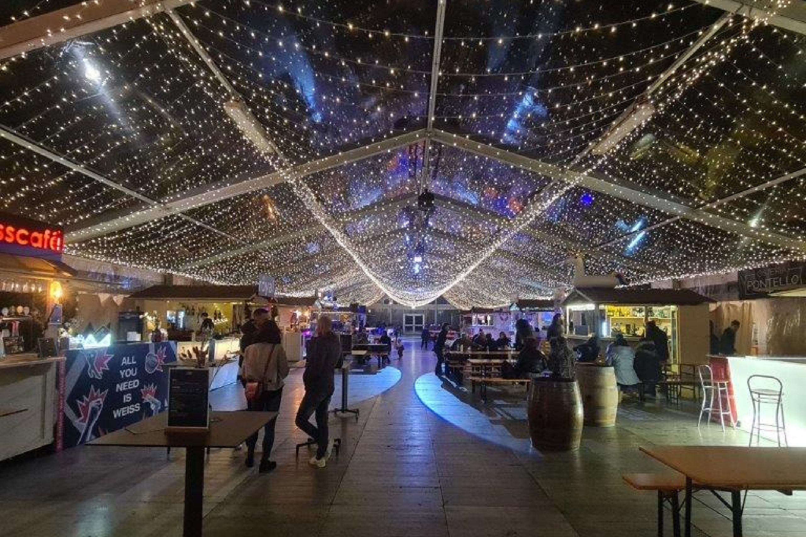 Un festoso mercato coperto con lucine in testa, bancarelle di cibo su entrambi i lati e persone sedute ai tavoli che camminano sotto una tenda trasparente di notte.