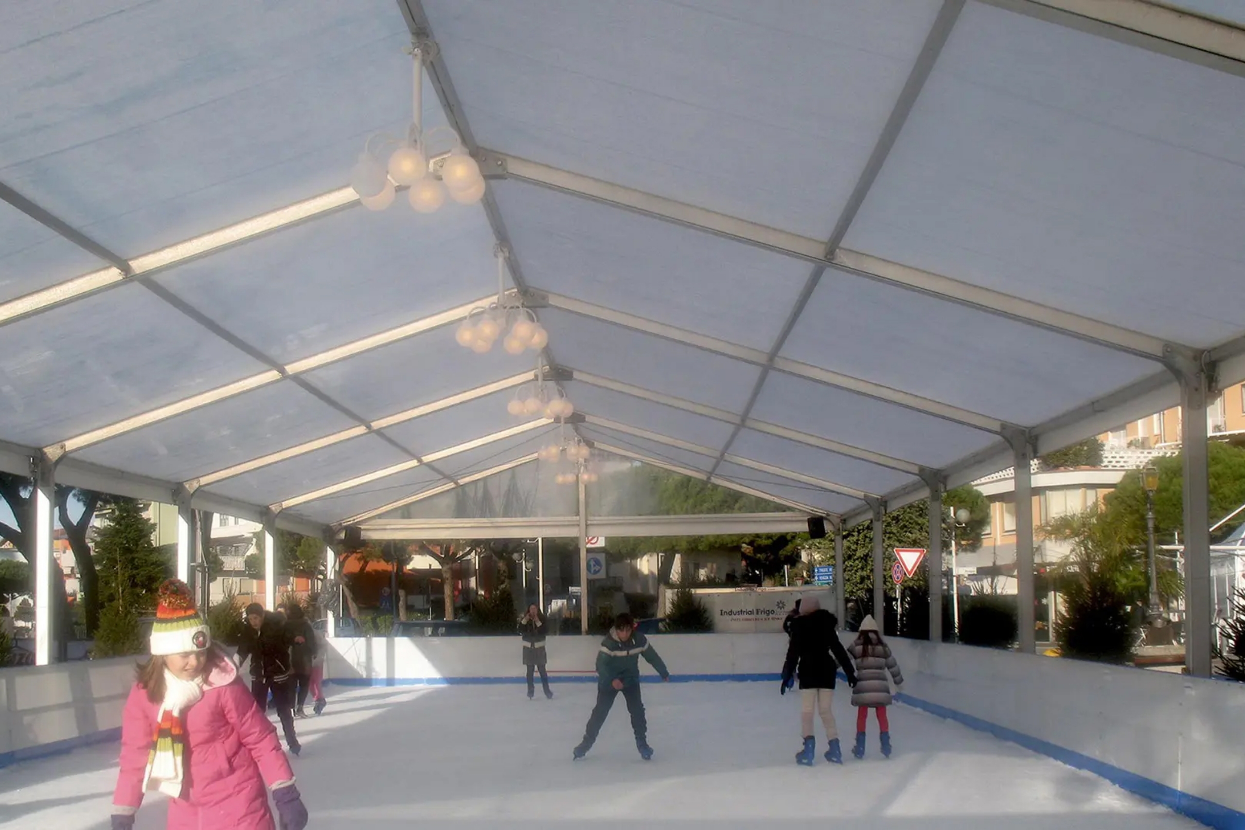 Persone che pattinano sul ghiaccio sotto una grande tenda bianca, con bambini e adulti che pattinano sulla pista. Alcuni indossano abiti invernali e caschi. Alberi ed edifici sono visibili sullo sfondo, oltre la tenda.