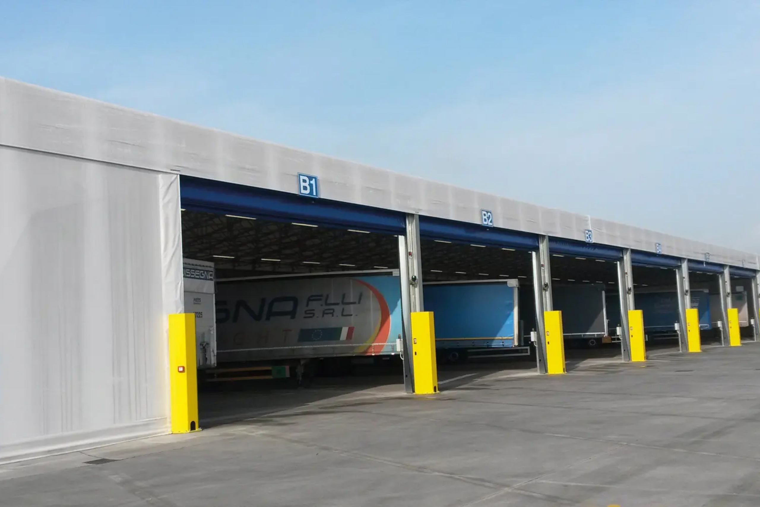 Una fila di banchine di carico con etichette blu (B1, B2, B3) sopra ogni baia, diversi camion parcheggiati nelle banchine e barriere gialle di fronte a ogni ingresso su una superficie pavimentata.