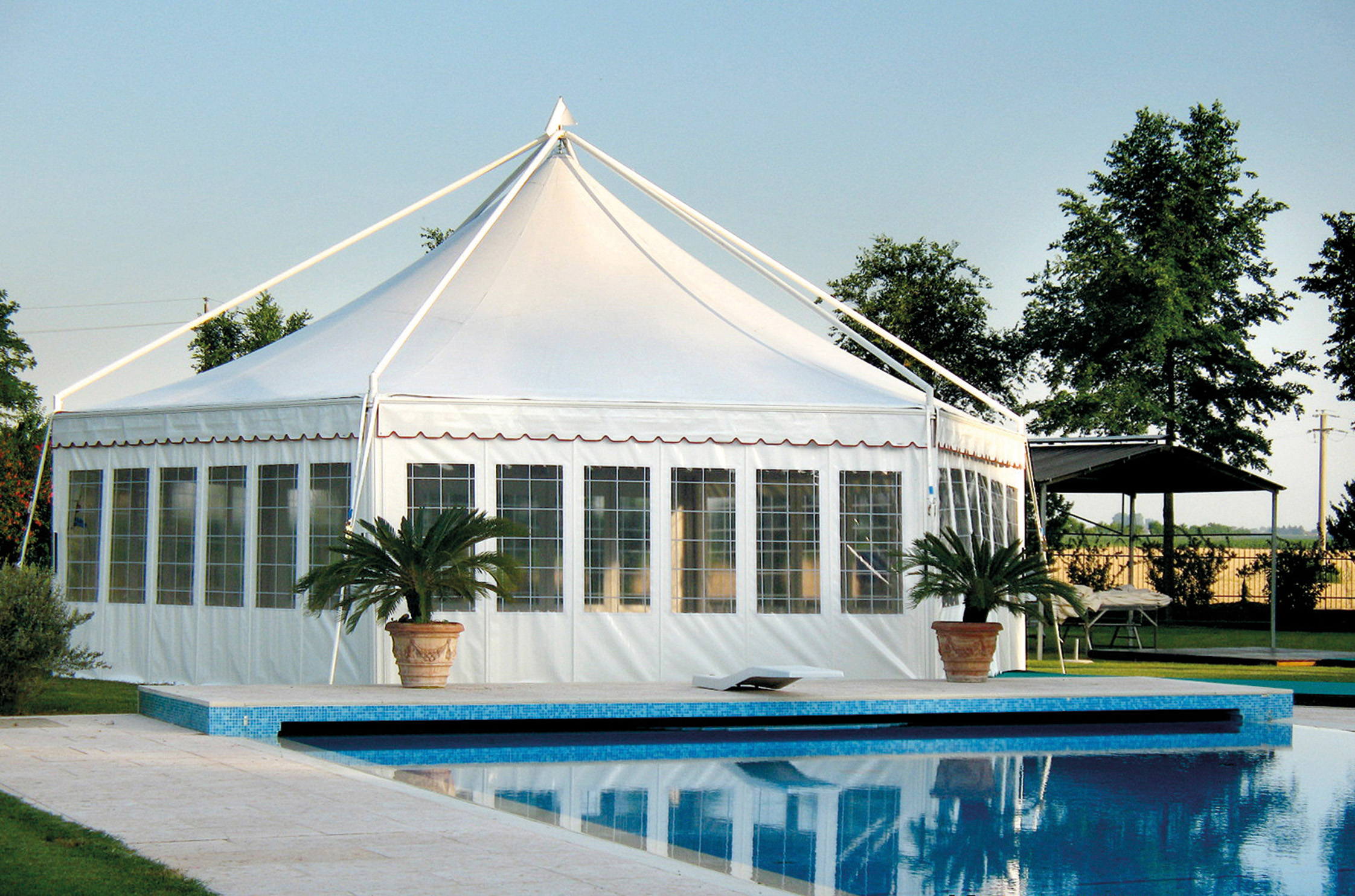 Nelle giornate limpide, accanto a una piscina blu, circondata dal verde e da palme in vaso, si erge un grande tendone bianco per eventi dotato di finestre.
