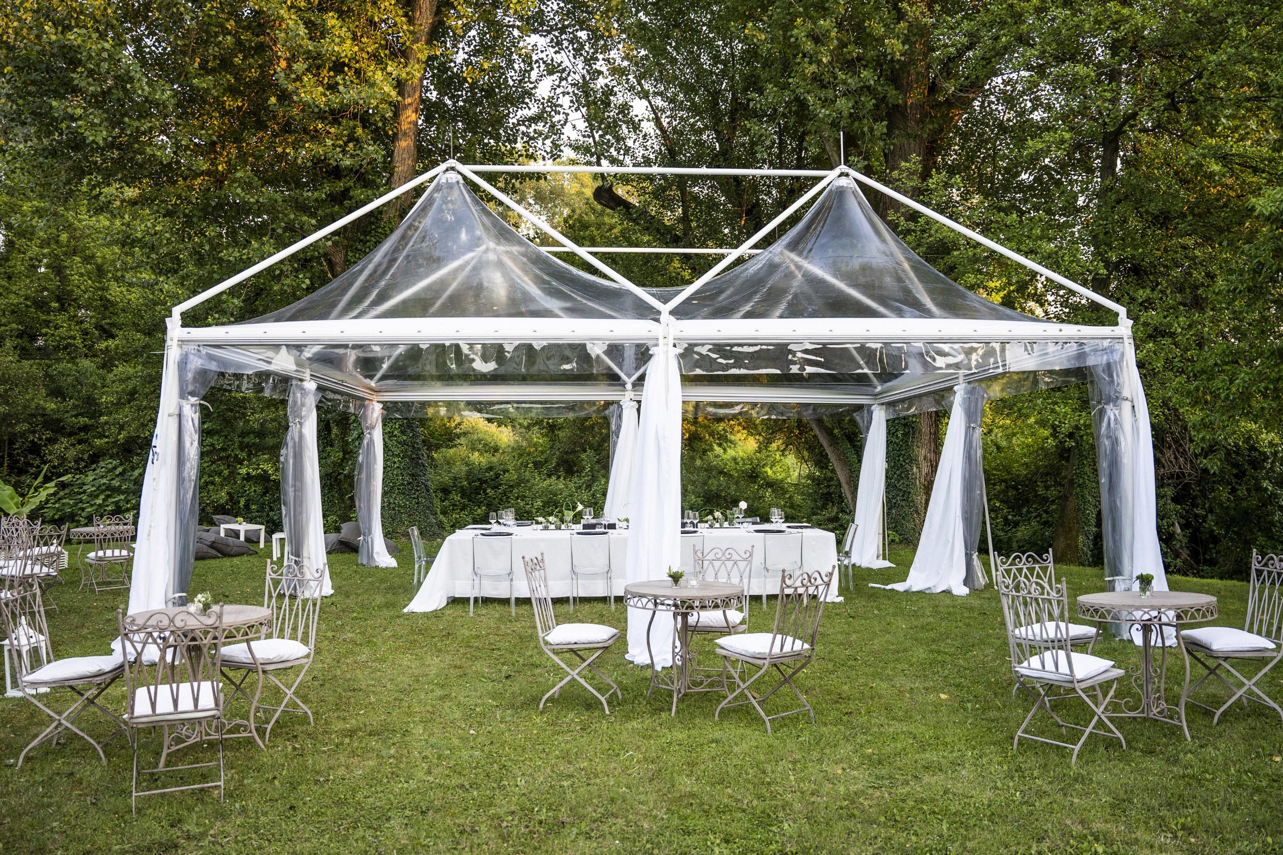 Una tenda trasparente con drappi bianchi si erge sull'erba, circondata da alberi. All'interno e intorno alla tenda ci sono diversi tavoli rotondi con sedie bianche, apparecchiati per un evento all'aperto. L'atmosfera è luminosa e tranquilla.