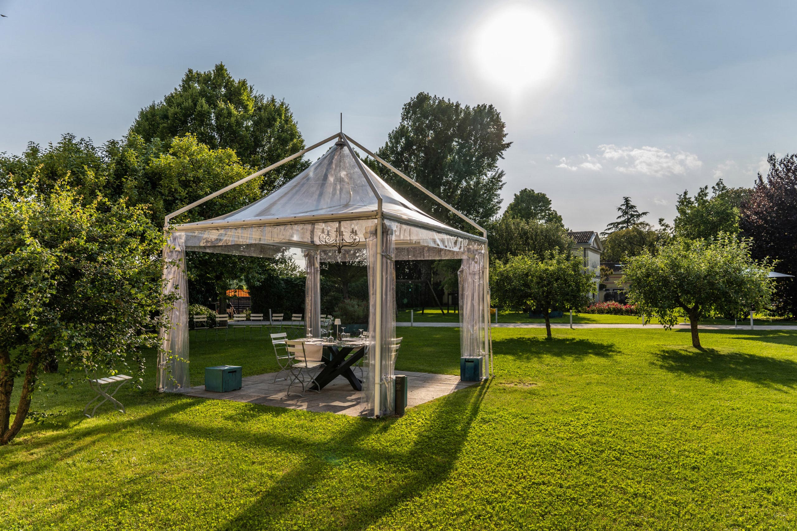 Una tenda trasparente è allestita su un prato verde circondato da alberi, con sedie pieghevoli, un tavolo e sgabelli blu all'interno. Il sole splende luminoso nel cielo azzurro.