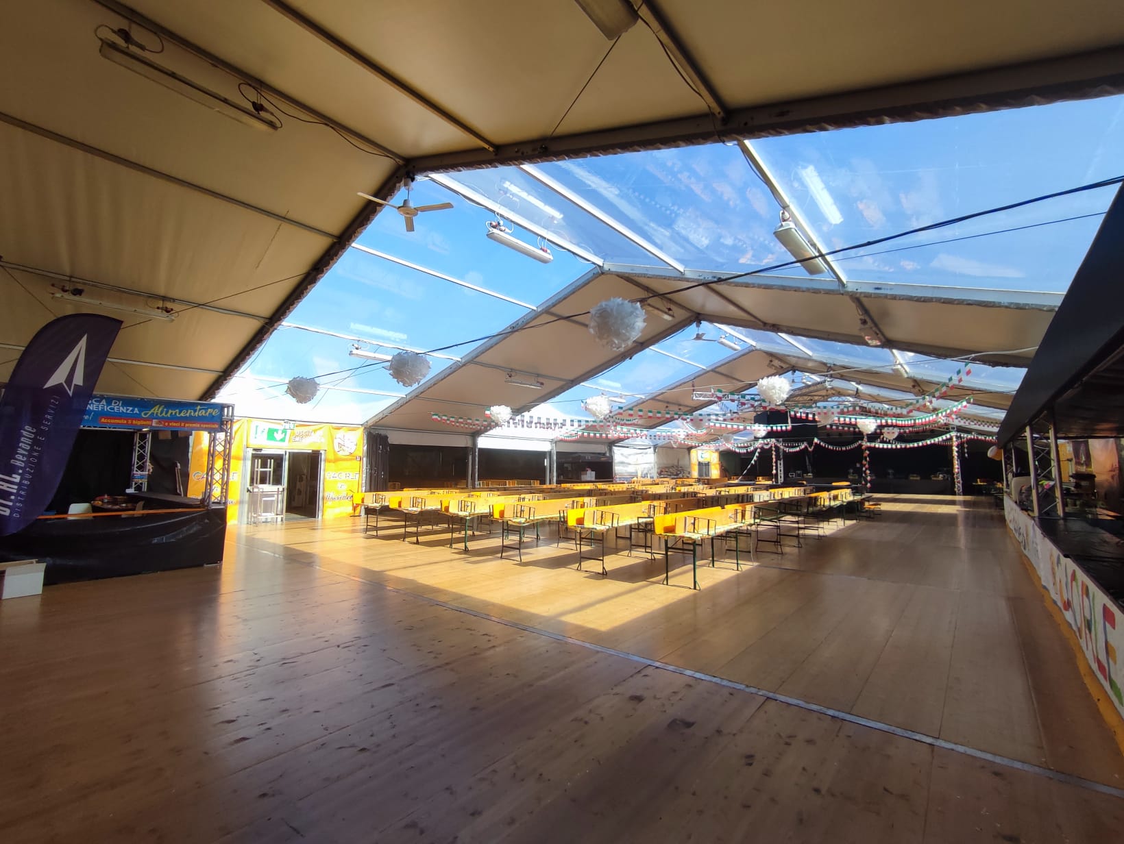 Interno di una grande tenda con pannelli trasparenti sul tetto, che lasciano filtrare la luce del sole. Lunghi tavoli e panche di legno sono disposti in file. Striscioni decorativi e lanterne pendono dal soffitto, creando un'atmosfera festosa.