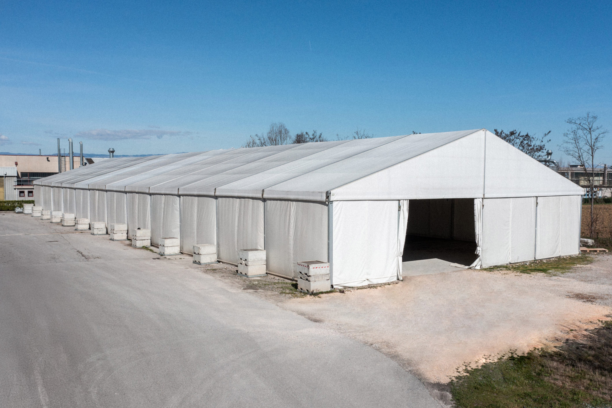 Una grande tenda temporanea bianca, composta da più sezioni, sorge su un'area pavimentata sotto un cielo azzurro e terso. La tenda è parzialmente aperta a un'estremità, con blocchi di cemento che ne rivestono la base.
