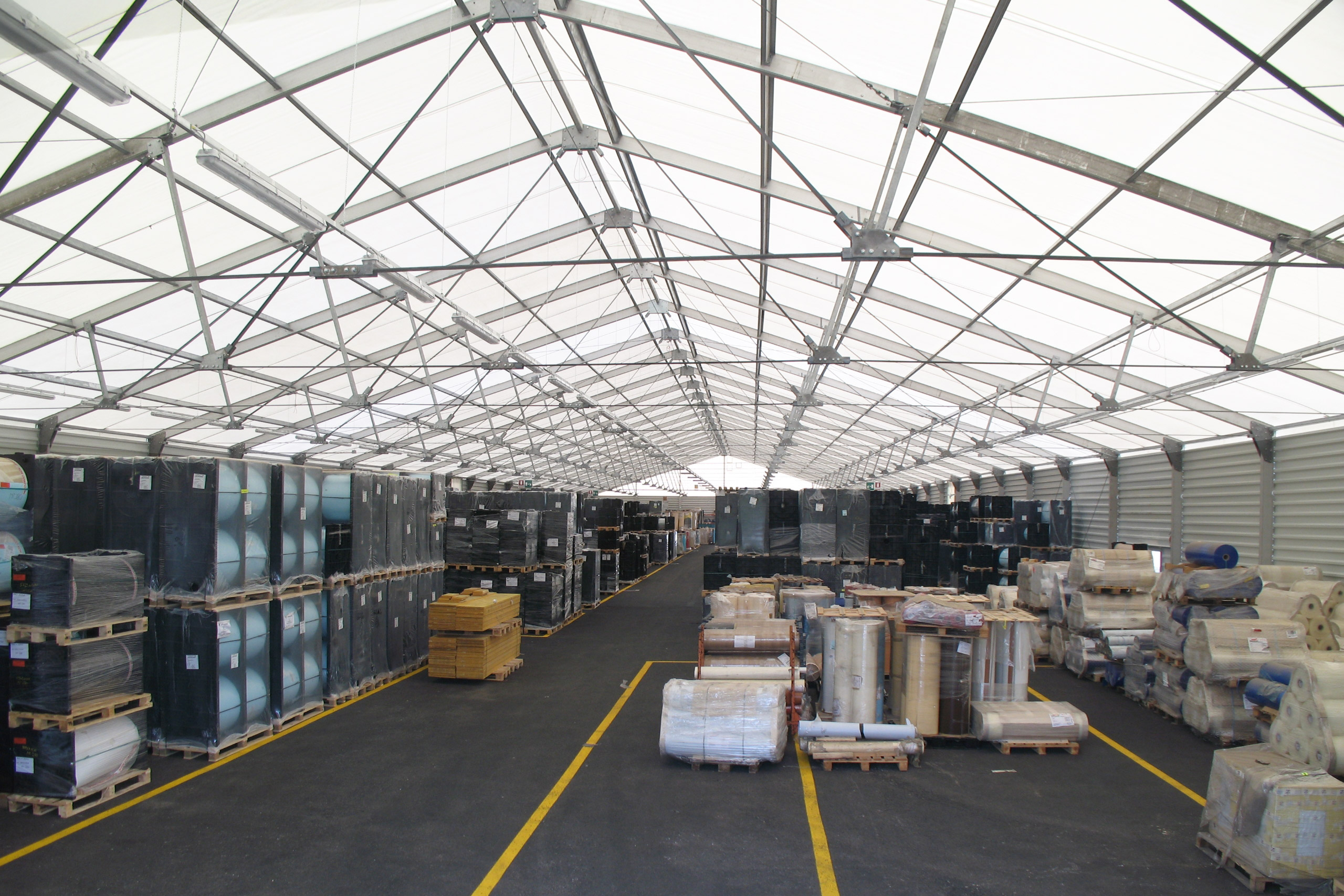 Un grande magazzino con un tetto traslucido, pieno di pallet di merci e materiali vari, ordinatamente disposti lungo entrambi i lati di un'ampia corsia centrale. Linee gialle segnano le corsie sul pavimento scuro.