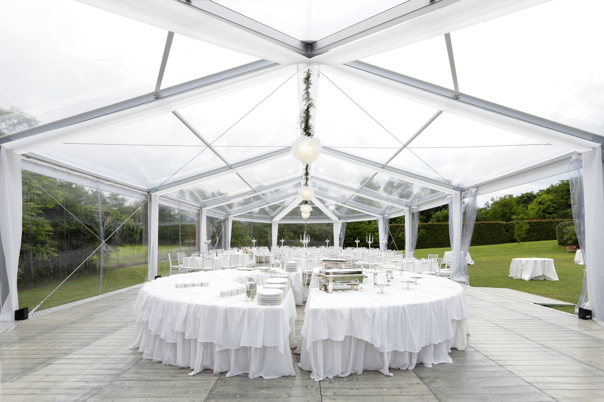 Una grande tenda trasparente allestita all'aperto con tavoli rotondi coperti da tovaglie bianche, eleganti tovaglie e sedie bianche, pronta per un evento formale o un matrimonio. Un verde lussureggiante circonda la location.