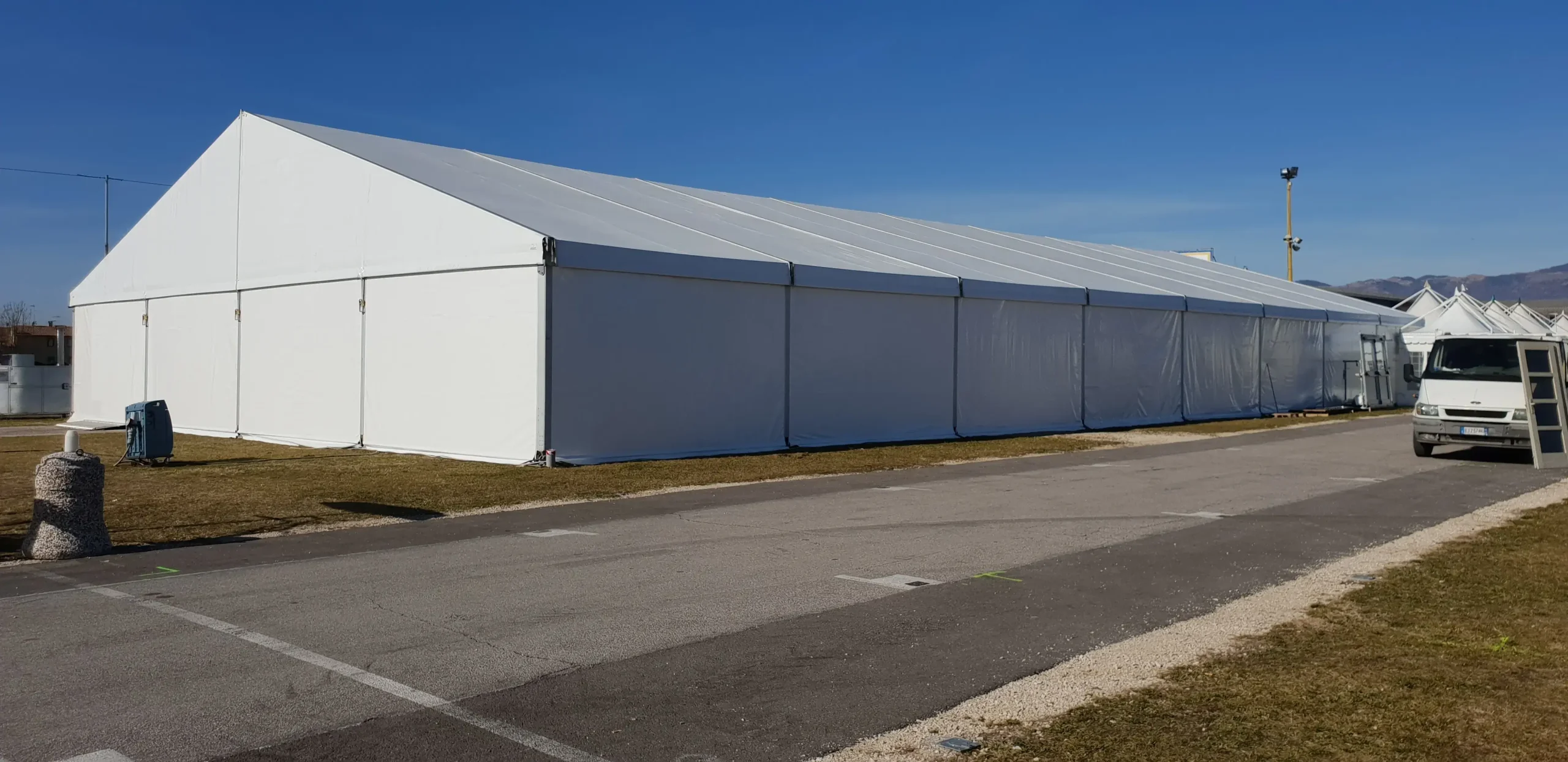 Una grande tenda bianca è montata su un prato accanto a una strada asfaltata, sotto un cielo azzurro e terso. Un piccolo camion è parcheggiato lì vicino e sullo sfondo si vedono le montagne.