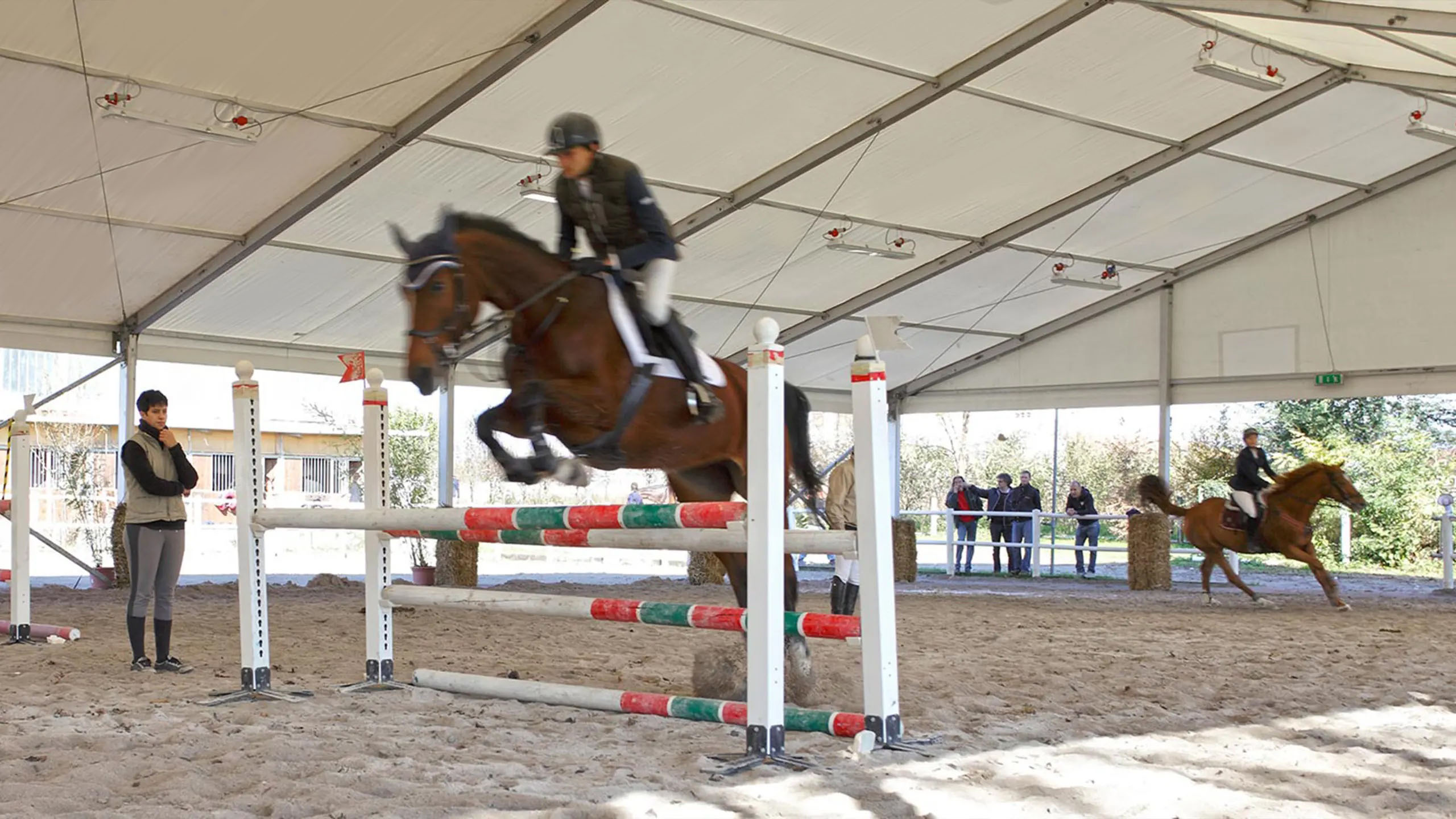 Un cavaliere in tenuta da equitazione sta saltando un ostacolo su un cavallo marrone in un'arena coperta. Nelle vicinanze, un altro cavallo e il suo cavaliere si stanno preparando. Gli spettatori osservano sullo sfondo.