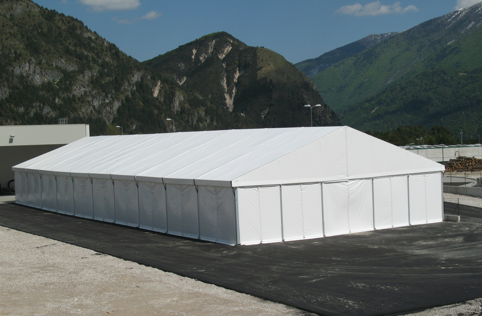 Un grande tendone bianco per eventi è posizionato su una superficie pavimentata, con montagne, colline verdi e un cielo parzialmente nuvoloso sullo sfondo.
