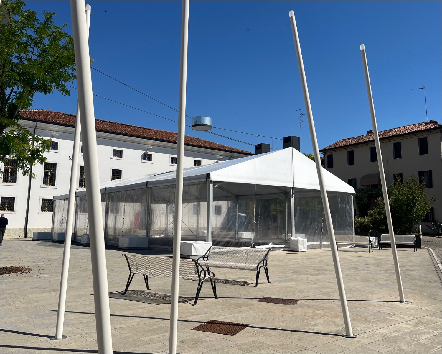 Una tenda bianca con i lati trasparenti si erge in un cortile pavimentato, circondata da moderni pali bianchi, panchine e alberi, con edifici a più piani sullo sfondo sotto un cielo azzurro e limpido.