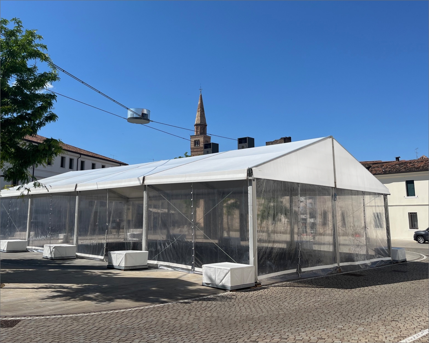 Una grande tenda bianca con i lati trasparenti è installata su un'area pavimentata sotto un cielo azzurro e terso, con il campanile di una chiesa e gli edifici circostanti visibili sullo sfondo.