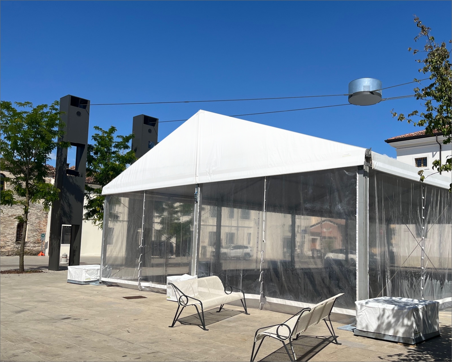 Una tenda bianca con i lati trasparenti si erge su una piazza pavimentata, circondata da panchine e alberi sotto un cielo azzurro e terso. Sullo sfondo si vedono edifici moderni e grandi strutture rettangolari.