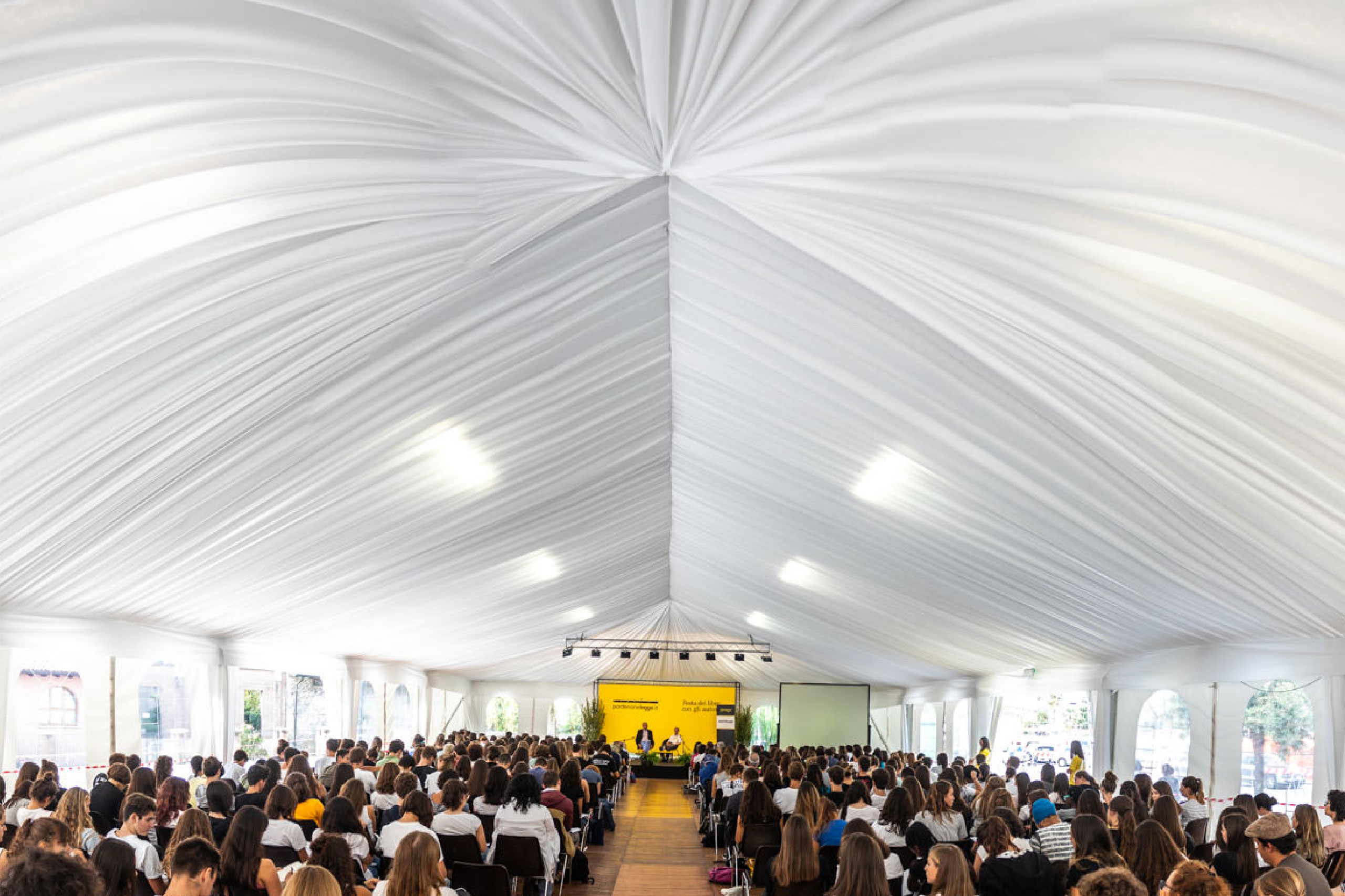Un folto pubblico siede sotto una tenda bianca con un telo drappeggiato, di fronte a un palco con relatori e uno sfondo giallo, durante una conferenza o un evento. Uno schermo per proiettore è visibile a destra del palco.