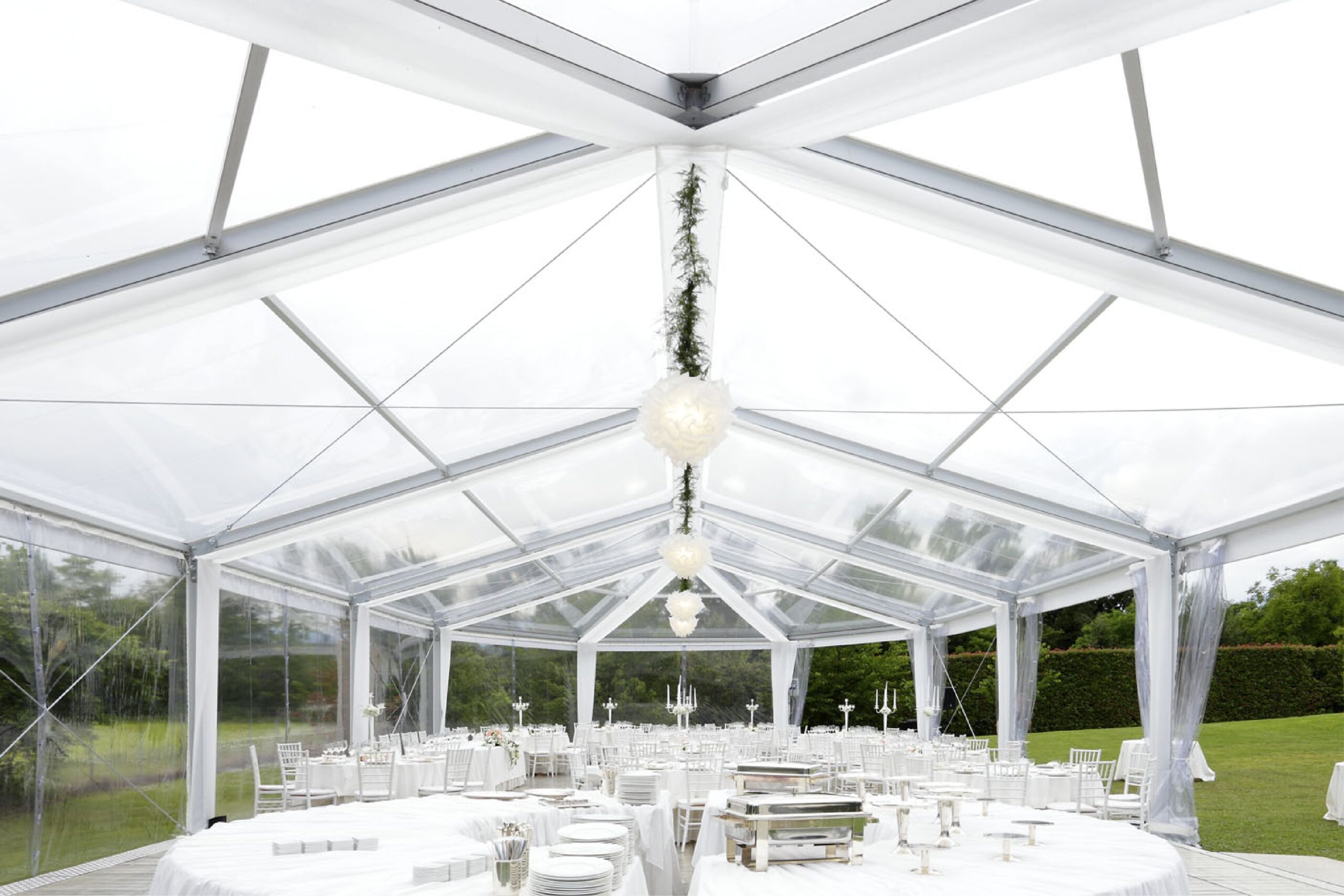 Un ricevimento di nozze in bianco sotto una tenda trasparente con tavoli rotondi coperti da teli bianchi, sedie bianche ed eleganti luci pendenti, circondati da una verde scenografia esterna.