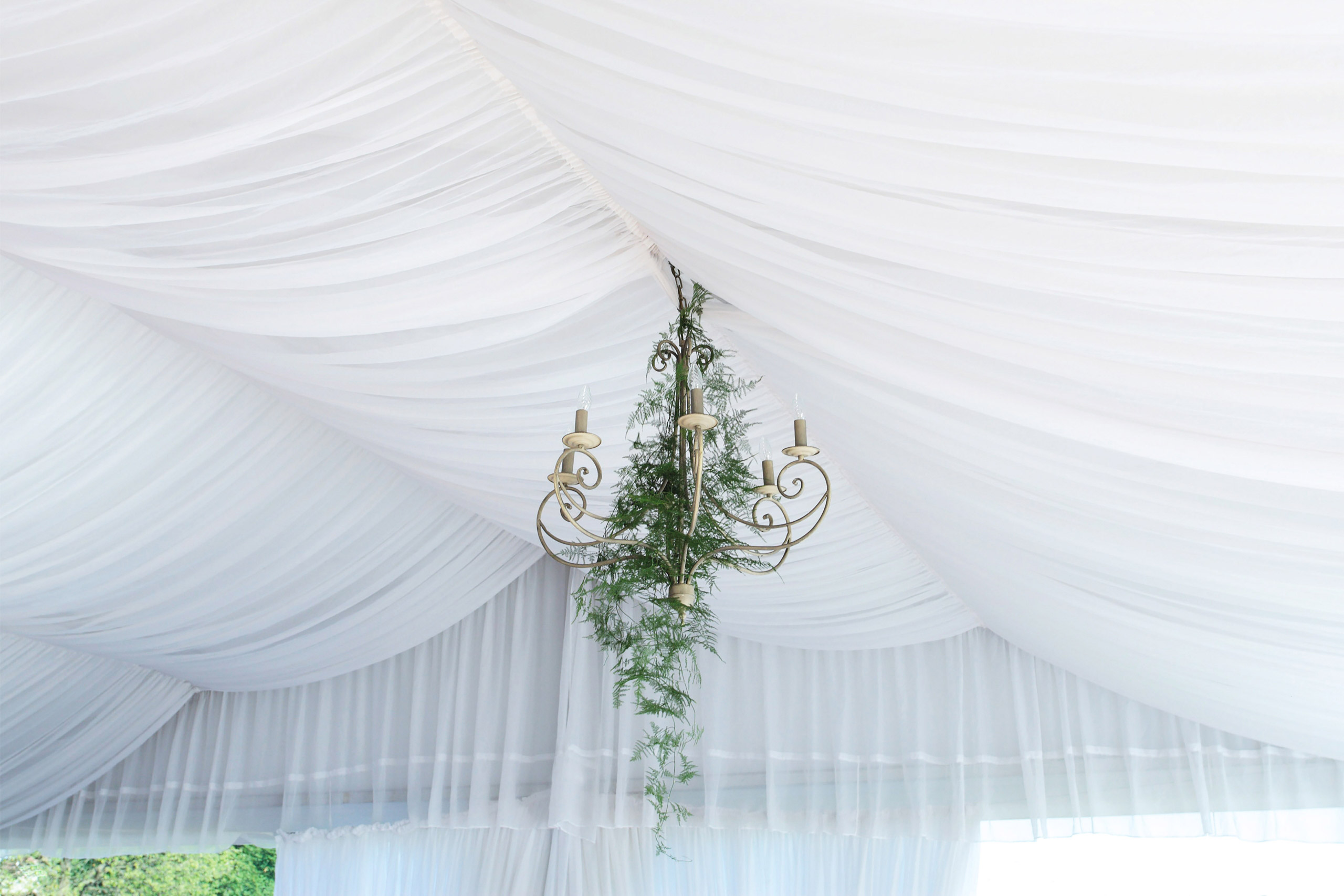 Un lampadario dorato decorato con elementi verdi pende da un soffitto drappeggiato di bianco, creando un'atmosfera elegante e ariosa.