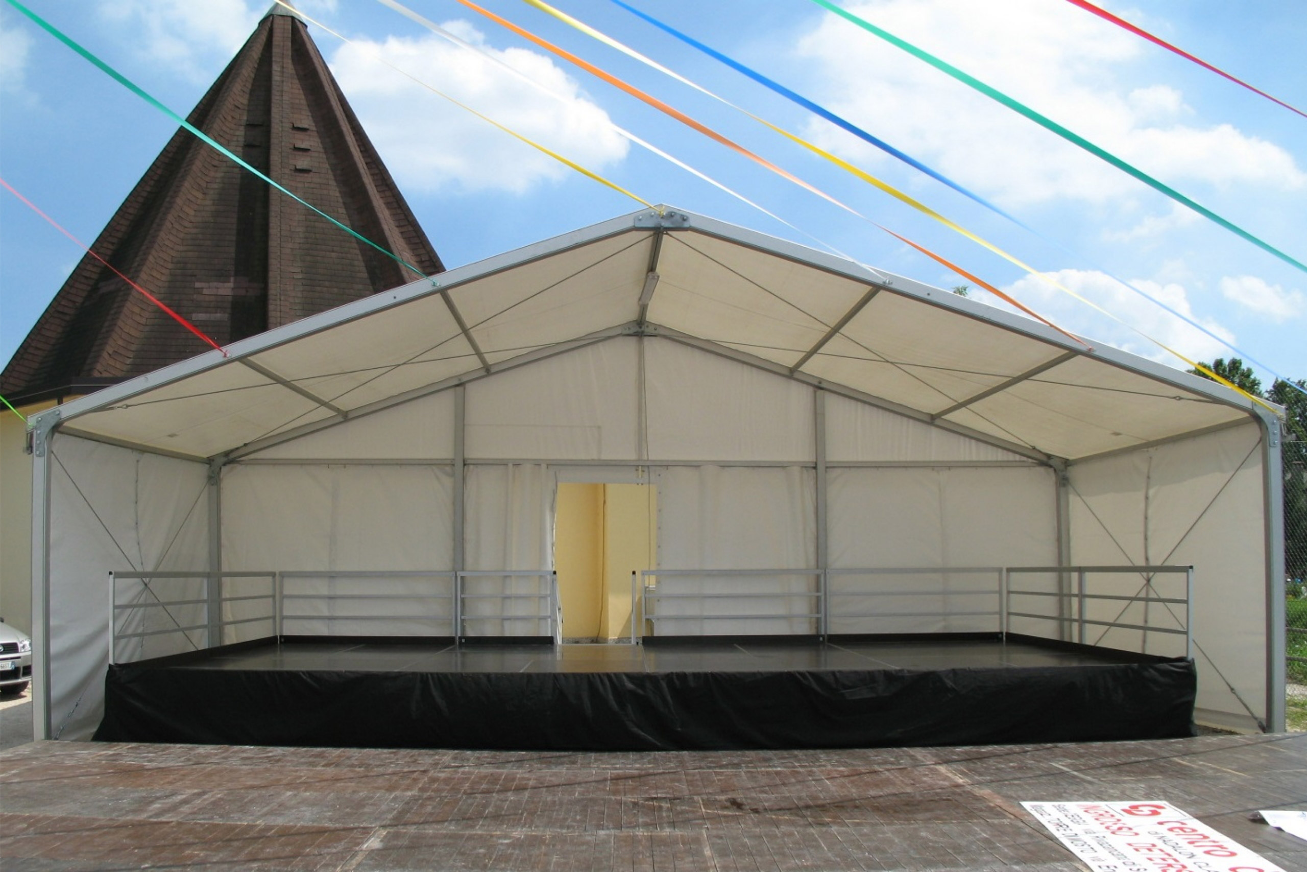Un grande tendone bianco per eventi all'aperto, aperto sul davanti e con un palco nero rialzato all'interno, allestito sul marciapiede, con festoni colorati in alto e un edificio triangolare in mattoni sullo sfondo.