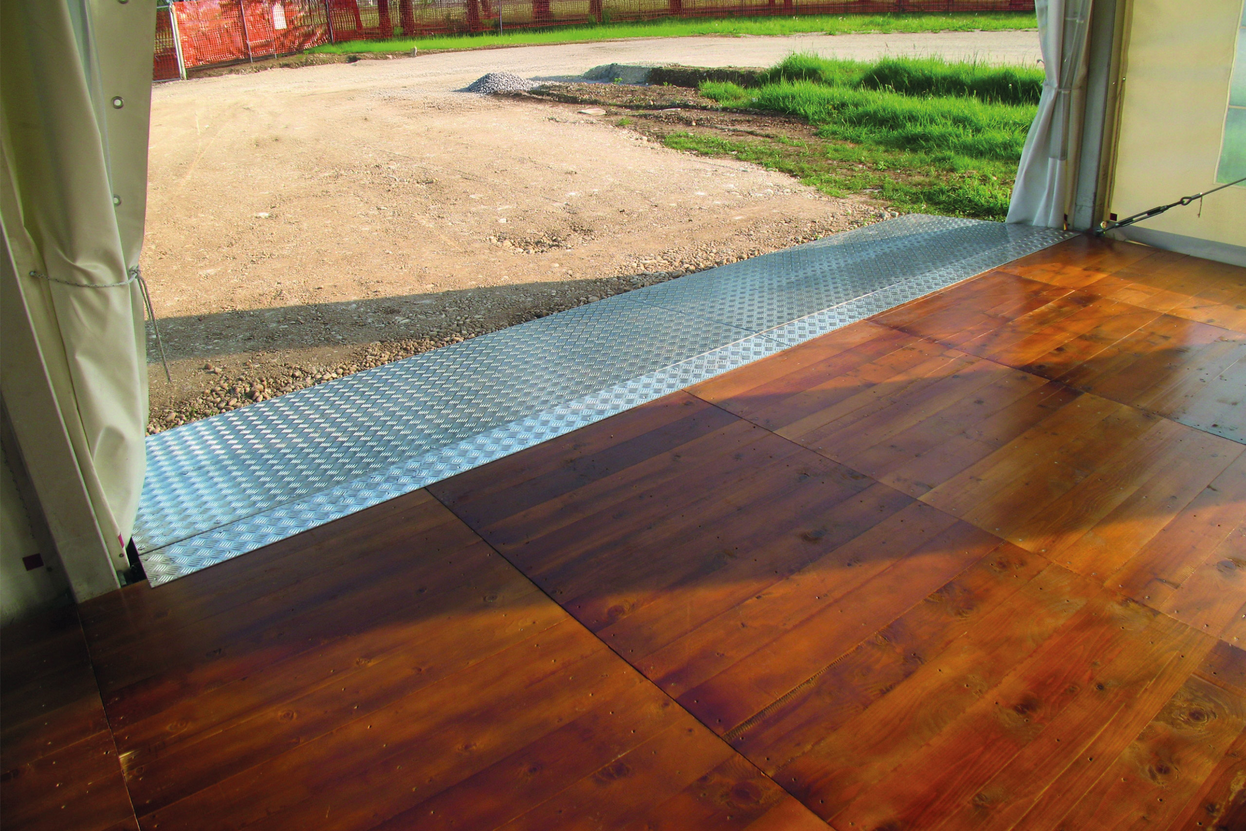 Vista dall'interno di una tenda, che mostra un pavimento in legno che conduce a una rampa metallica e a una strada sterrata esterna. Sullo sfondo si vedono erba verde e una recinzione da cantiere.