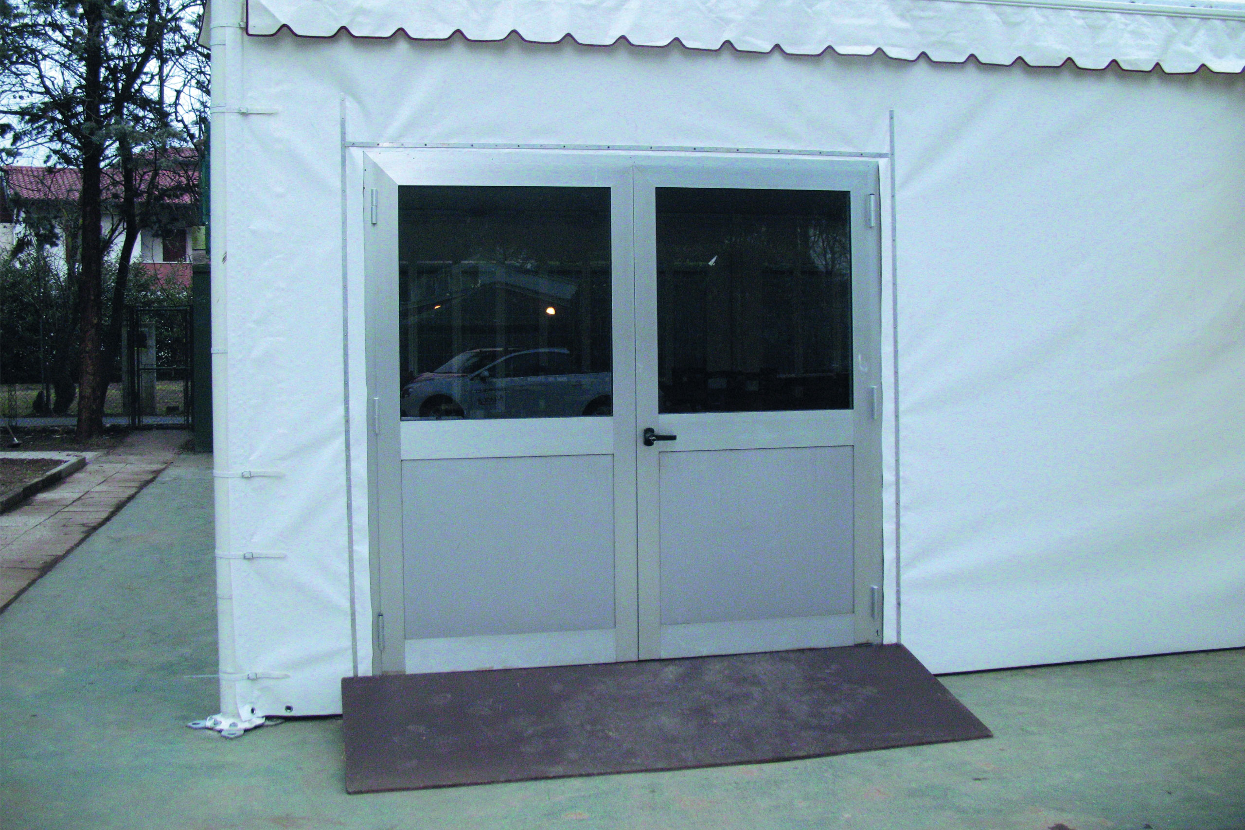 Una porta a doppia anta con telaio bianco è inserita in una struttura bianca a forma di tenda. Davanti alle porte, c'è una rampa marrone per l'accesso alle sedie a rotelle. Sullo sfondo si vedono alberi e un'auto parcheggiata.