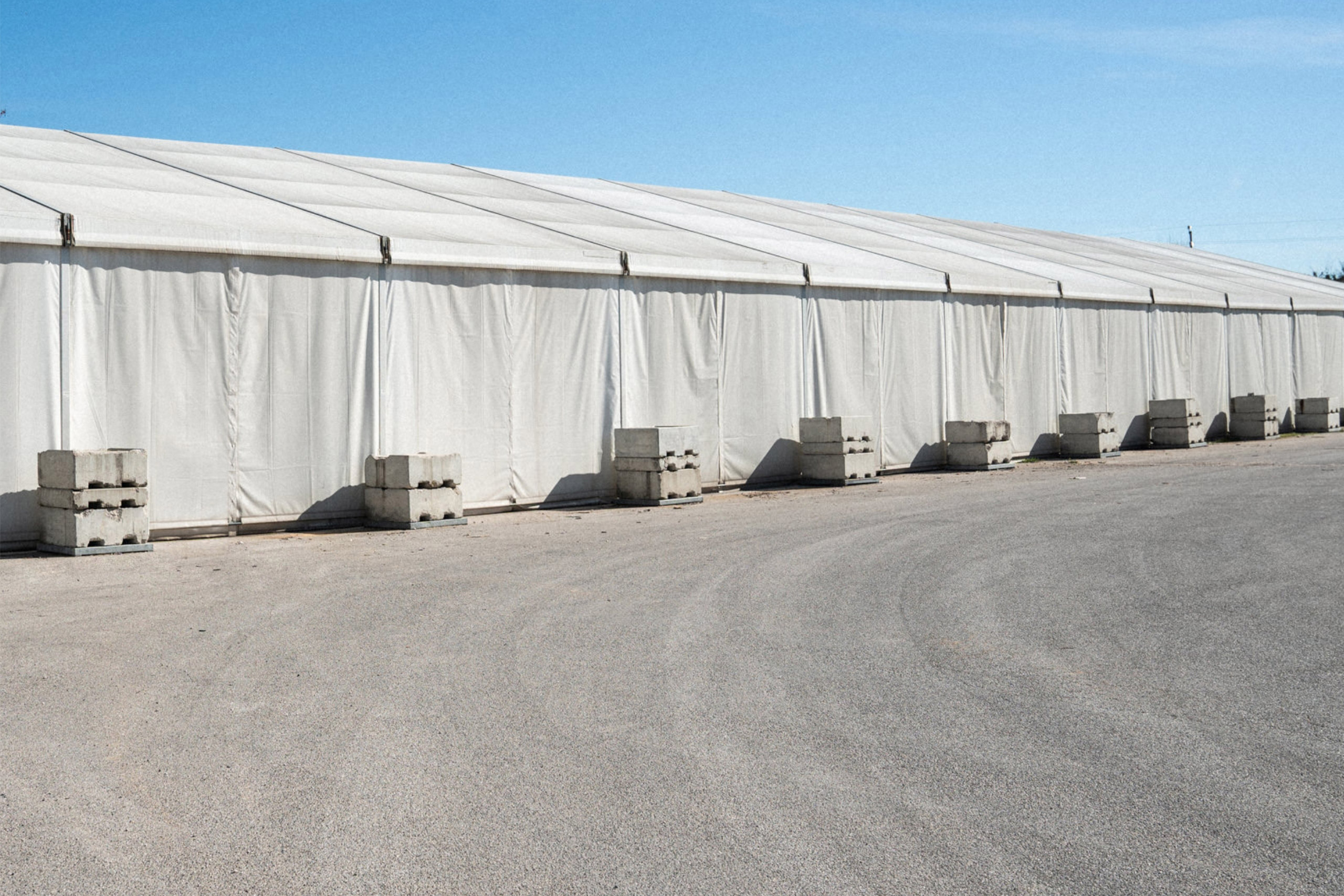 Una grande tenda bianca con i lati chiusi è ancorata tramite blocchi di cemento ed è installata su un'area pavimentata vuota sotto un cielo azzurro e terso.