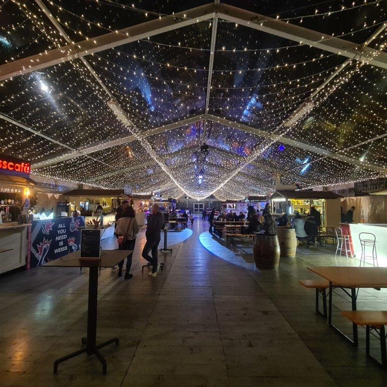 Un grande mercato coperto o un'area gastronomica con un soffitto trasparente ricoperto di lucine, che creano un effetto stellato. Le persone passeggiano e si radunano intorno a bancarelle e tavoli, in un'atmosfera festosa e vivace.
